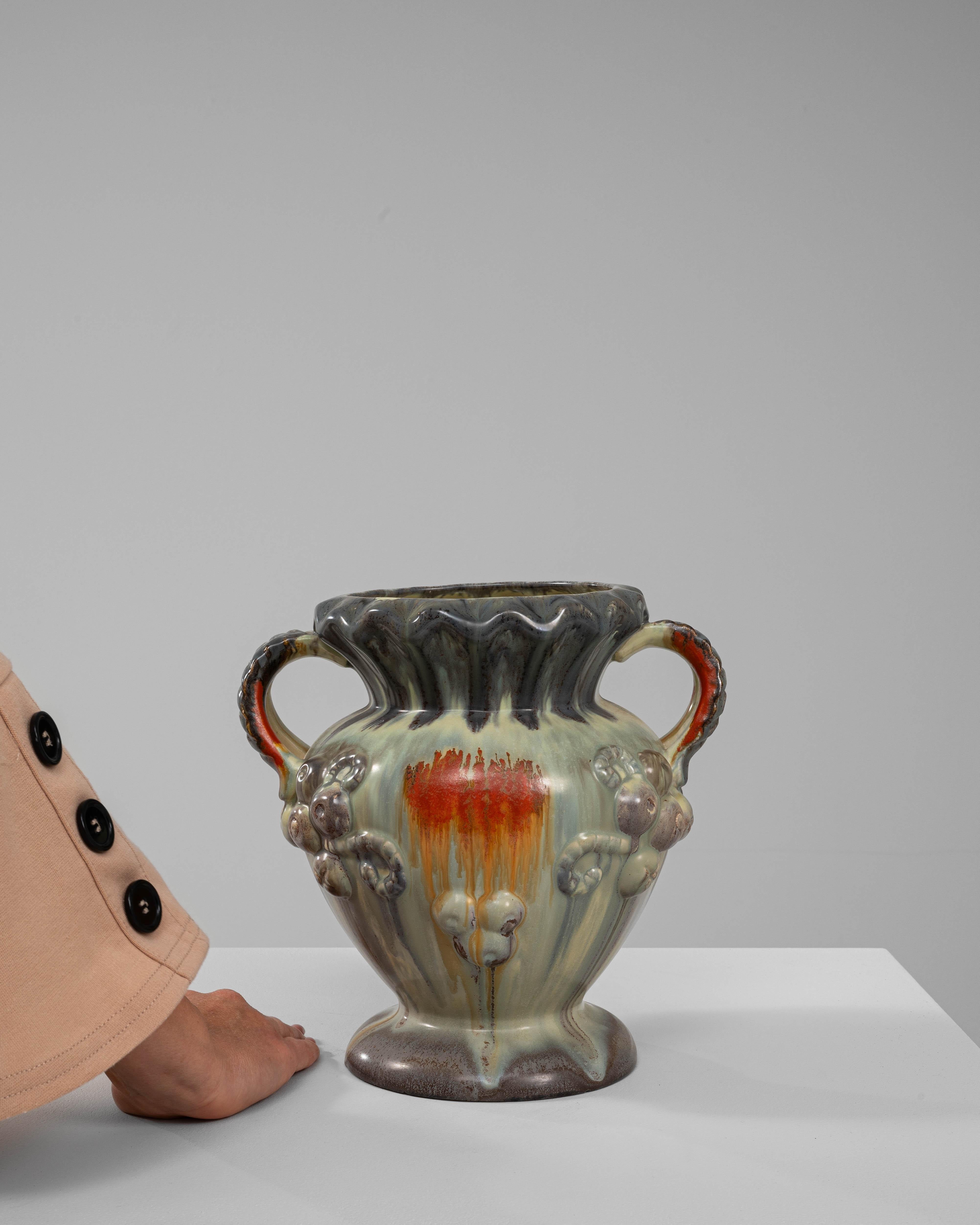 Questo vaso in ceramica belga del XX secolo emana eleganza e fascino grazie alla sua lavorazione artigianale. Caratterizzato da un bordo svasato e smerlato e da due manici ornati da intricati dettagli intrecciati, il vaso combina funzionalità e