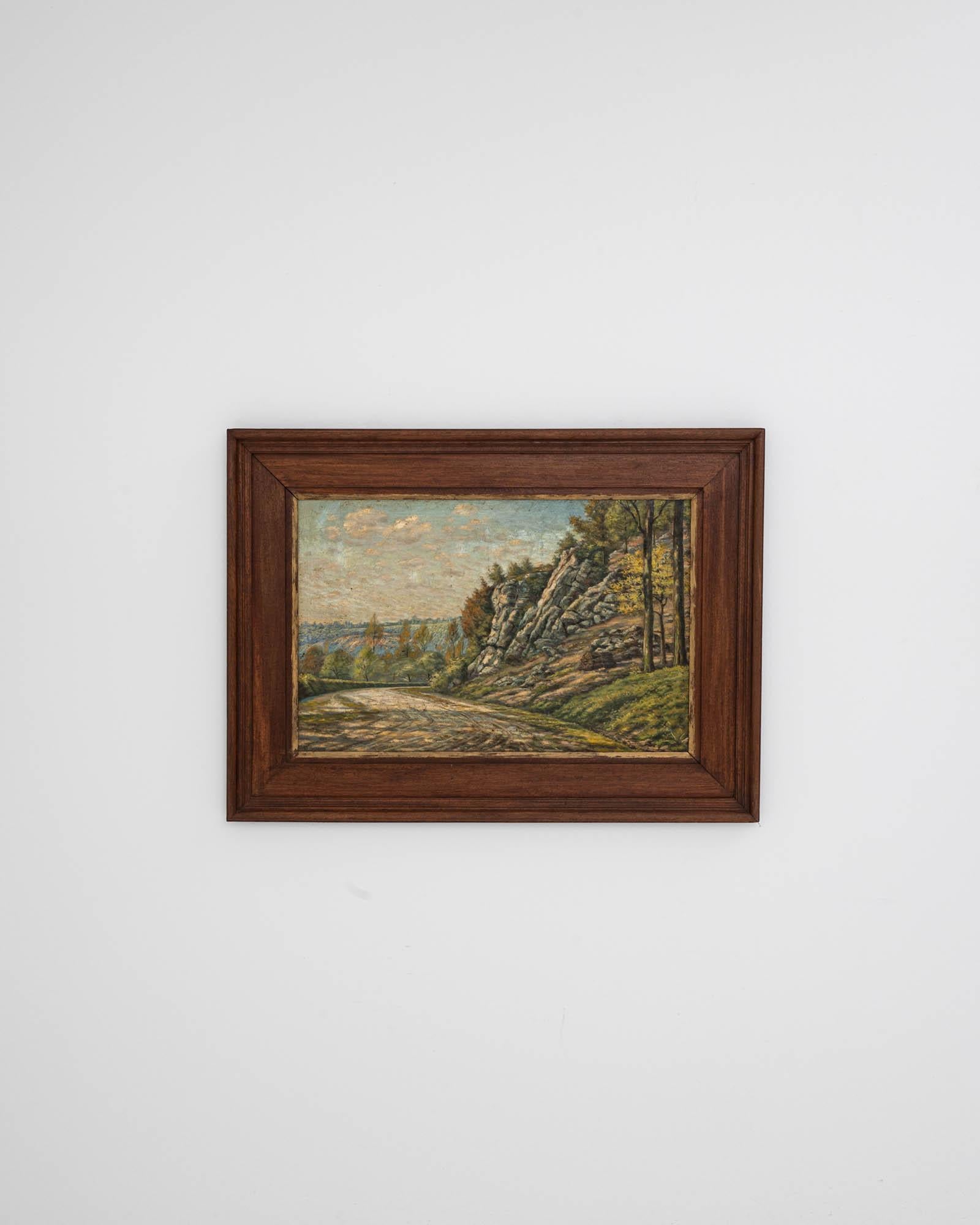 Inscrite dans un élégant cadre en bois de grande taille, cette peinture du XXe siècle représente un paysage paisible avec une route de village ouverte qui serpente à travers de vastes champs et des arbres, probablement en Belgique, le lieu d'origine