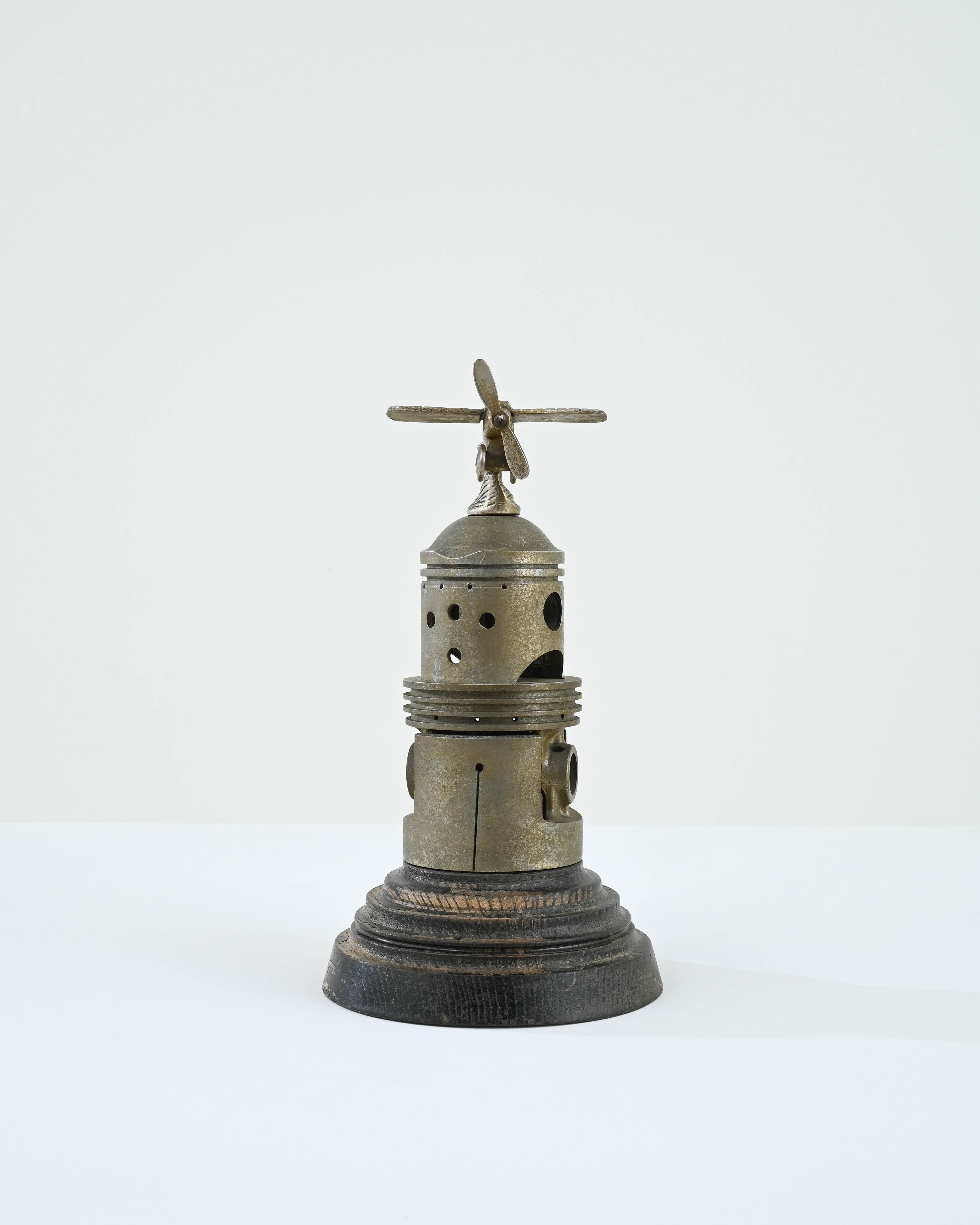A metal decoration created in 20th century Belgium. A propellered plane rests above a unique structure, seemingly created from repurposed engineering parts, resembling a futuristic tower. With richly patinated alloyed metal resting upon a round,