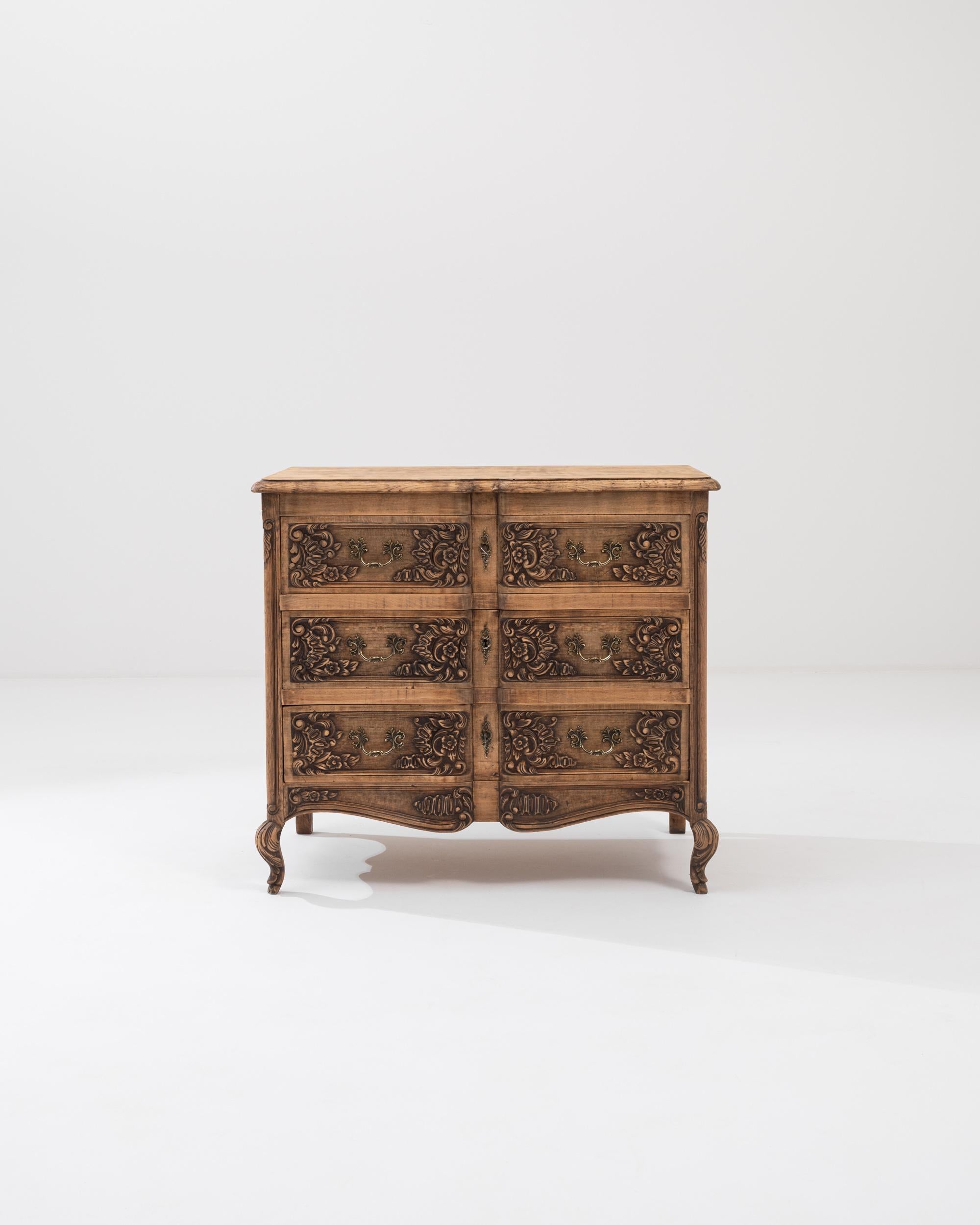 A wooden chest of drawers from 20th century Belgium. This ornate chest of drawers is lavishly decorated with swirling floral patterns that wreath around its drawer handles and curved feet. Tasteful accents such as brass drawer handles and keyholes