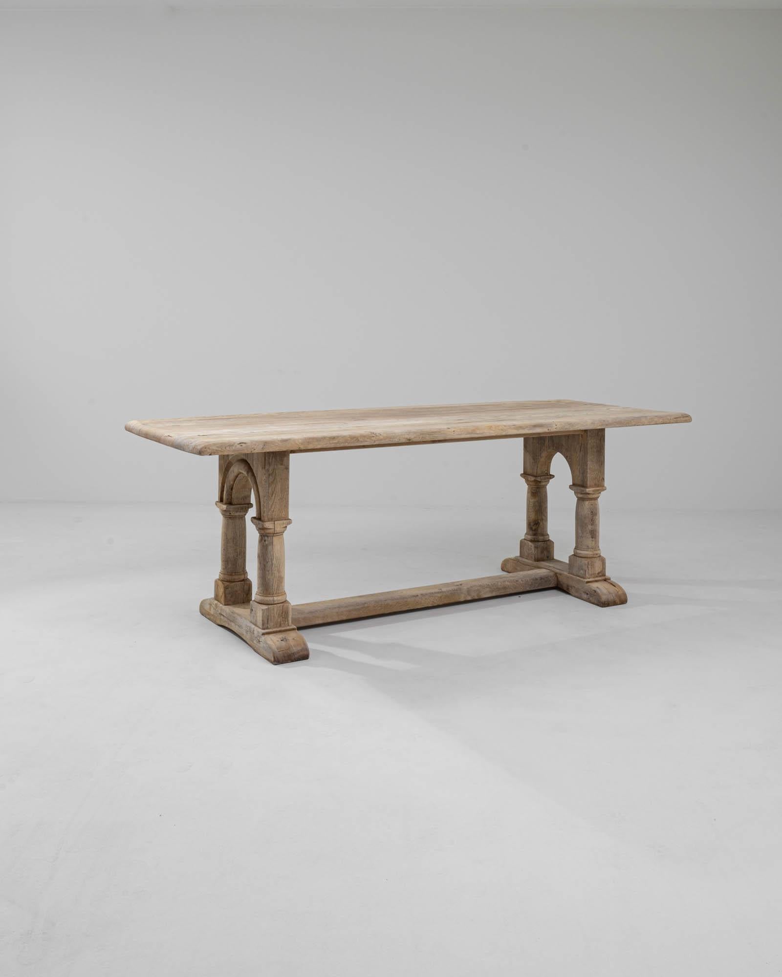 A wooden dining table made in 20th century Belgium. This stout and sturdily constructed trestle table is composed of two pairs of legs who stand like columns, connected by towering arches. A delicate bleaching process has spread across its surface