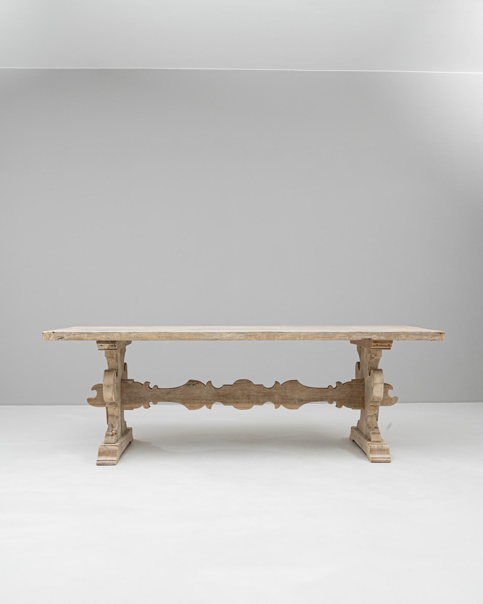 This 20th-century Belgian oak dining table is a captivating blend of artistry and functionality. Its robust construction features a sculptural trestle base with intricate scrollwork and a beautifully carved stretcher for added visual appeal and
