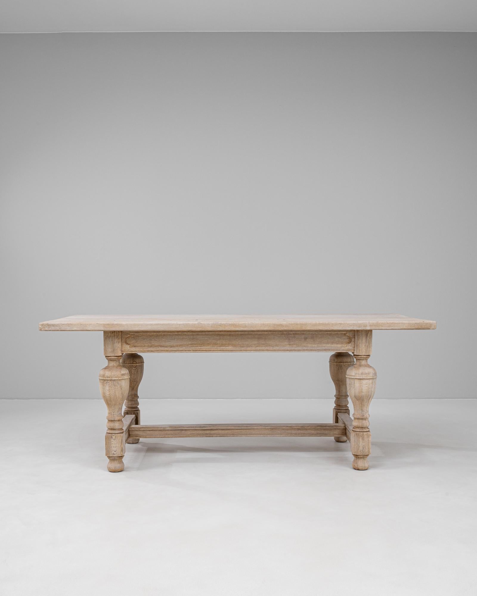 Mesa de comedor de roble belga del siglo XX, con tablero rectangular sostenido por robustas patas torneadas en balaustre unidas por un montante en H. El acabado en roble claro y natural resalta el bello veteado y la sutil pátina de la madera,