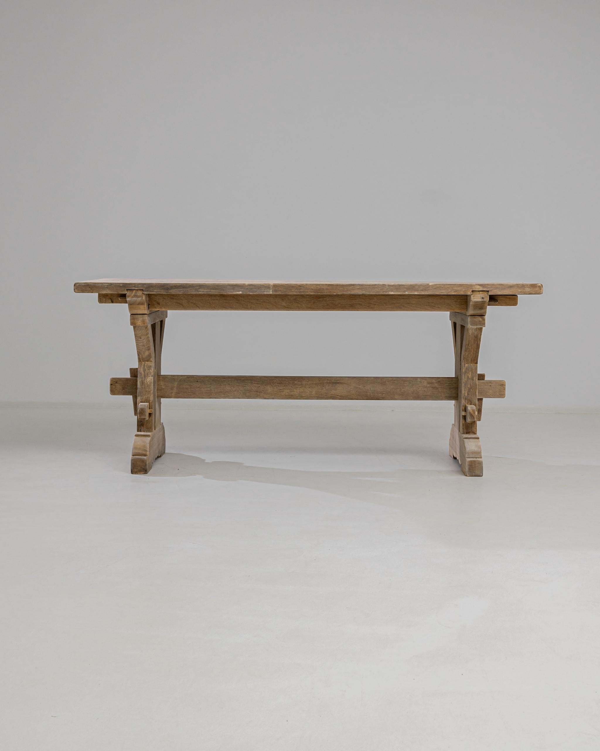 A country dining table in natural oak from 20th Century Belgium. Trestle legs, joined by a stretcher, create a farmhouse silhouette. Visible wooden pegs and rustic mortise and tenon joints showcase the beauty of traditional craftsmanship. The dusky