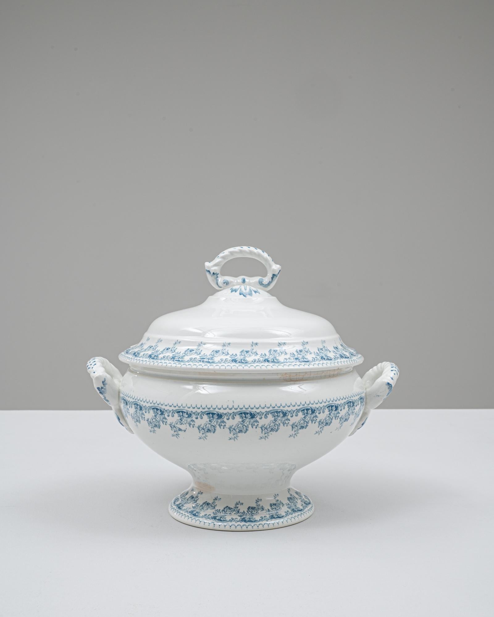 Cette soupière en porcelaine belge du 20e siècle, édition spéciale, présente un mélange élégant de formes classiques et de détails décoratifs raffinés. Sa silhouette gracieuse est rehaussée par une base sur piédestal et des poignées évasées, chacune