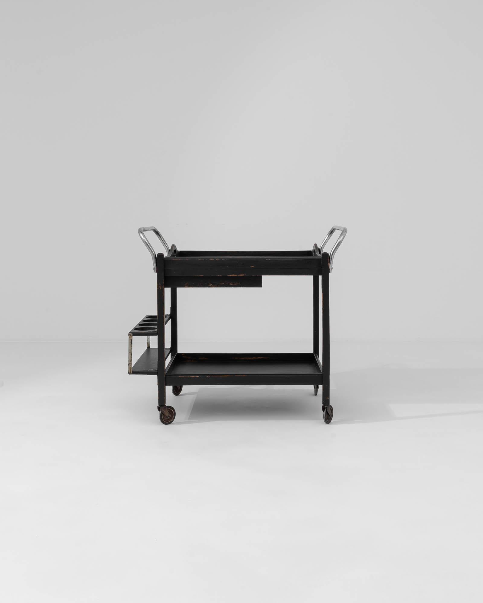 Step back in time with this 20th-century wood black patinated bar cart on wheels, an authentic piece of history designed to elevate your home entertainment experience. Crafted with care, its sturdy wooden frame is enhanced by a rich black patina