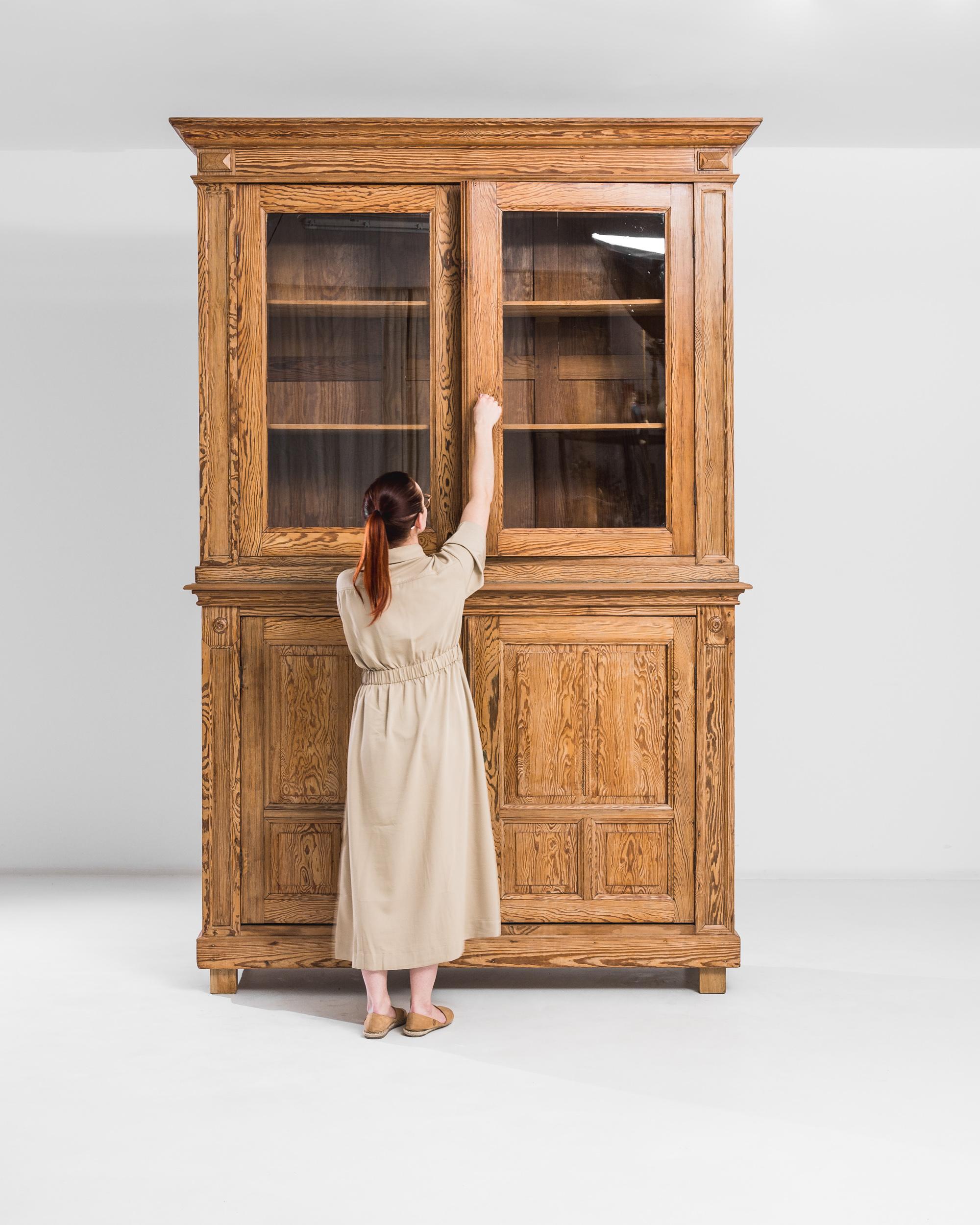Embrace the charm of a 20th Century Belgian Wooden Cabinet, exuding warmth and sophistication through its inviting finish. This remarkable piece features two glass doors, offering a glimpse into well-appointed shelves on the upper tier. Below, two