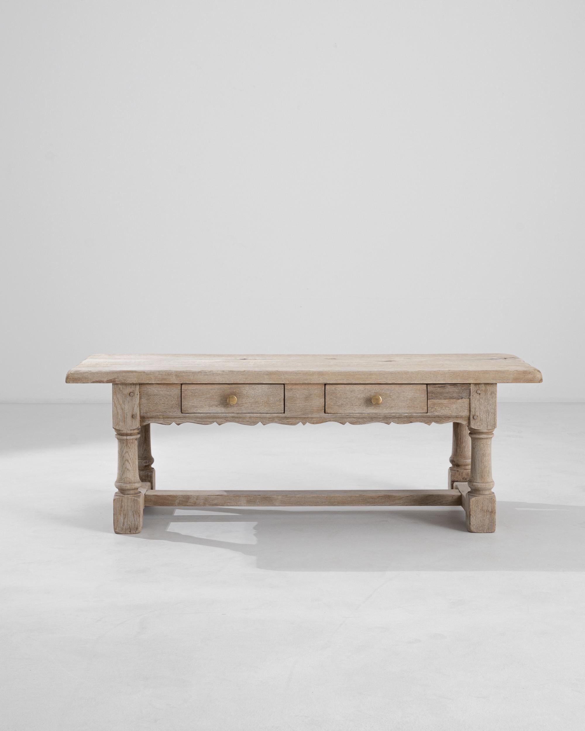 A bleached oak Belgian coffee table from the 20th century. In addition to an expansive table top, this coffee table also contains two large sliding drawers. Lathed legs, brass hardware, wooden pins, and hand-carved apron patterning all speak to the