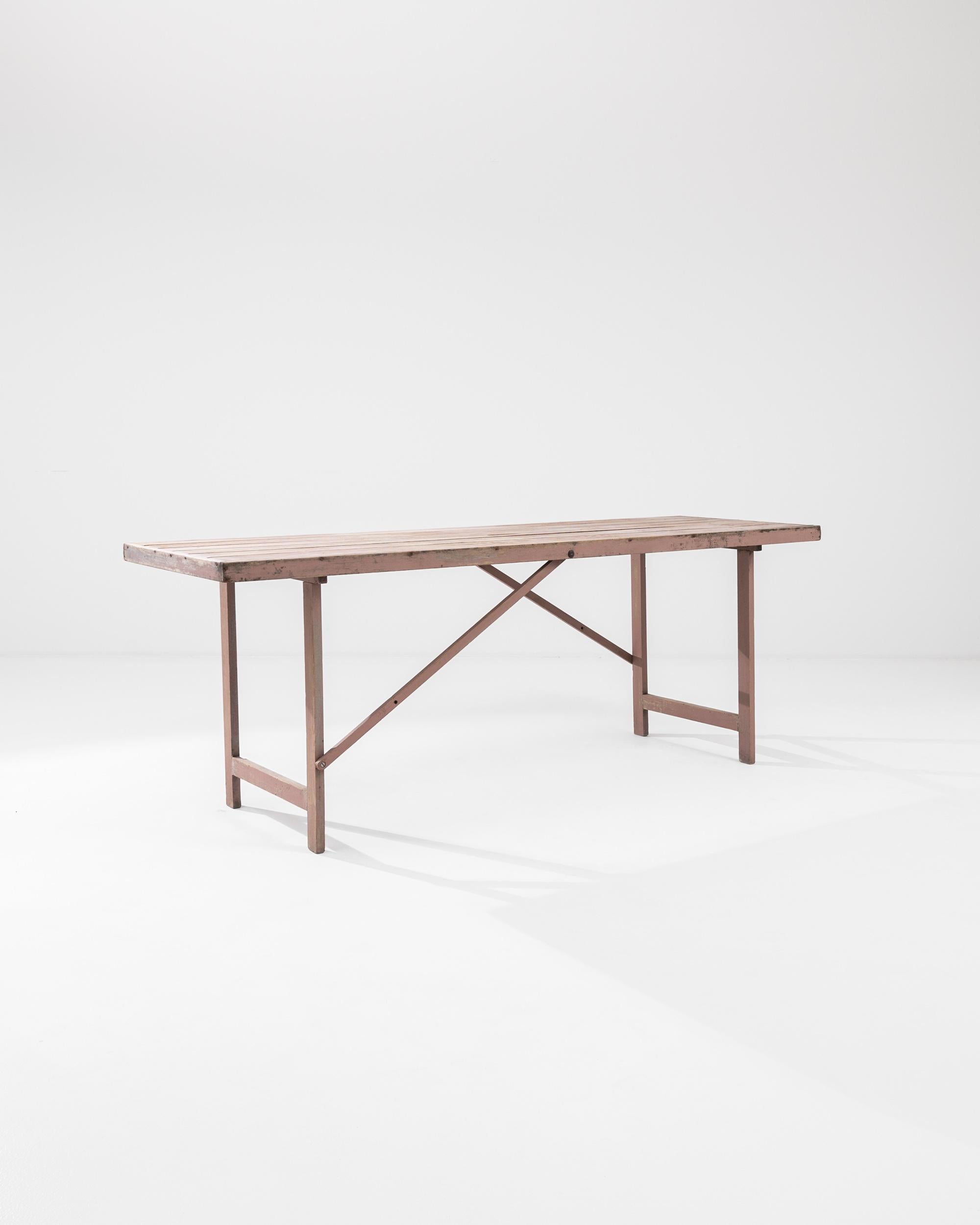 A wooden table made in 20th century, Belgium. The simple yet intriguing structure of this table features two countervailing diagonal supports which provide a pleasing geometric accent to its design. The rose colored pastel paint that coats its