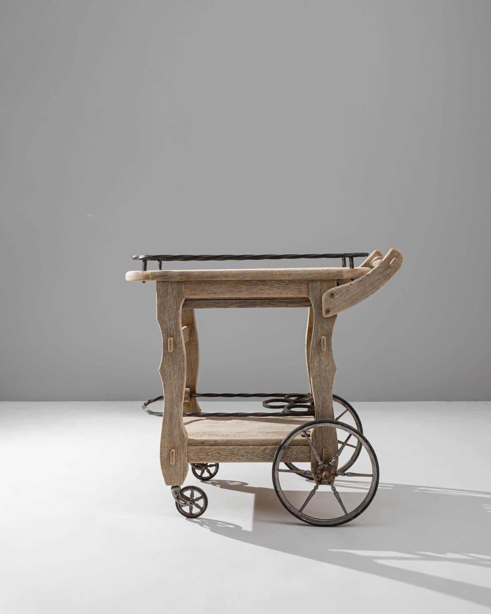 A wooden bar cart created in 20th century Belgium. Boasting a rustic farmhouse charm, this portable bar cart achieves a delightful beauty through its playful shape and metal fixtures. With subtly carved legs and twisted wrought iron details, the