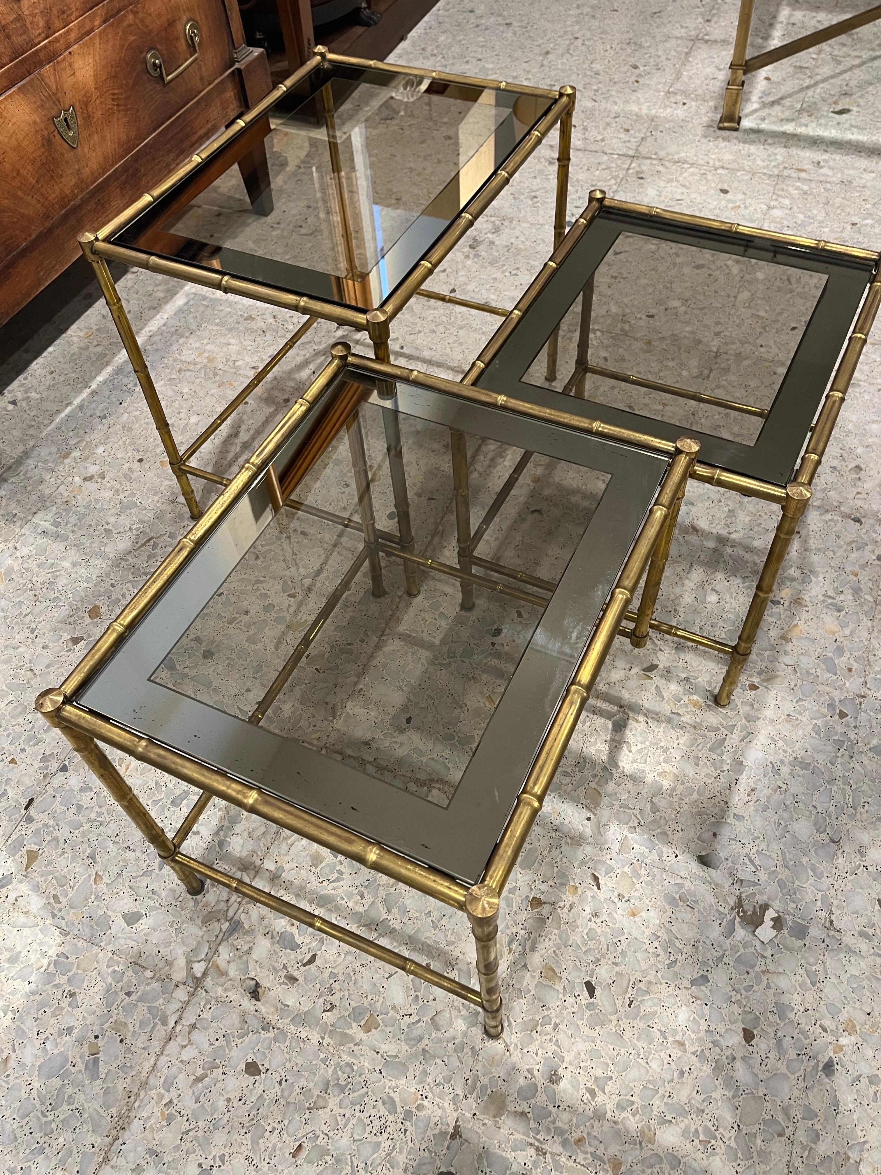 A nesting tables, composed of three tables of different sizes, imitating bamboo, and made of brass. The table top is two-tone glass, a gray tone framing the top. 
This type of table was very very emblematic among others, of the great French house of