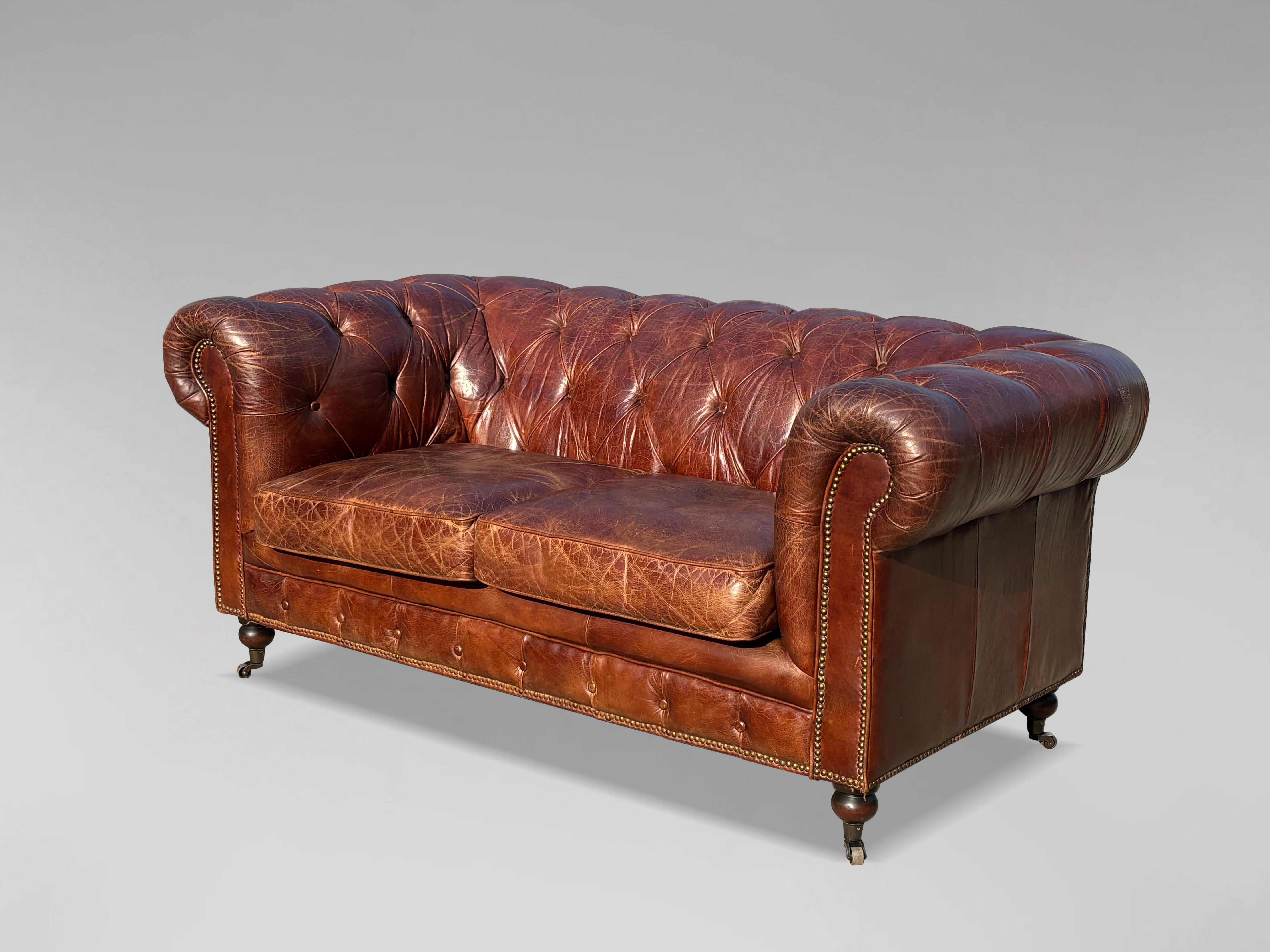 Chesterfield de dos plazas de cuero marrón del siglo XX, de buena calidad, con dos cojines sueltos, elevado sobre patas torneadas que terminan en ruedas. Con remates de latón tachonados a mano individualmente. Un buen ejemplo de un cómodo