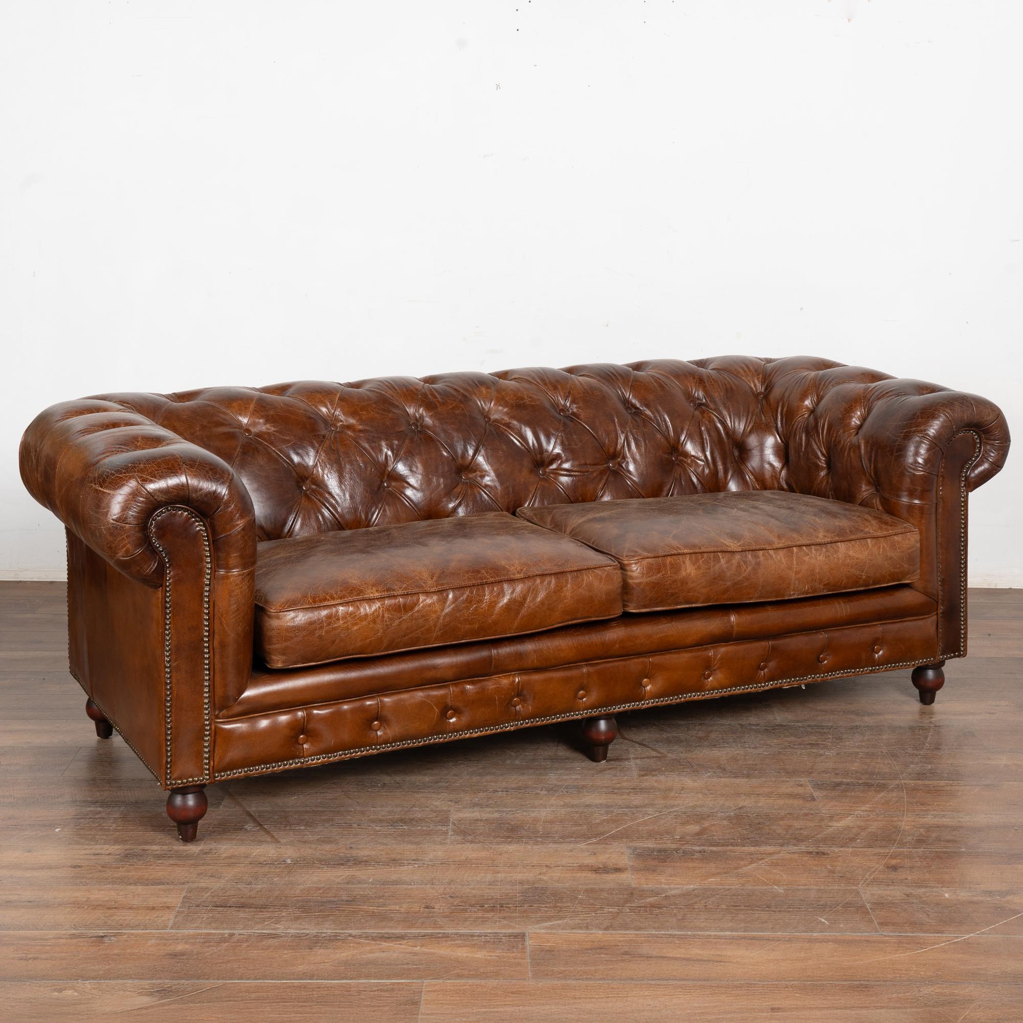 Das reiche braune Vintage-Leder zieht Ihre Aufmerksamkeit auf dieses wunderbare Chesterfield 7' lange Zweisitzer-Sofa.
Die traditionelle, getuftete und geknöpfte Rückenlehne, die gerollten Armlehnen und die mit Nägeln verzierten Akzente sorgen für