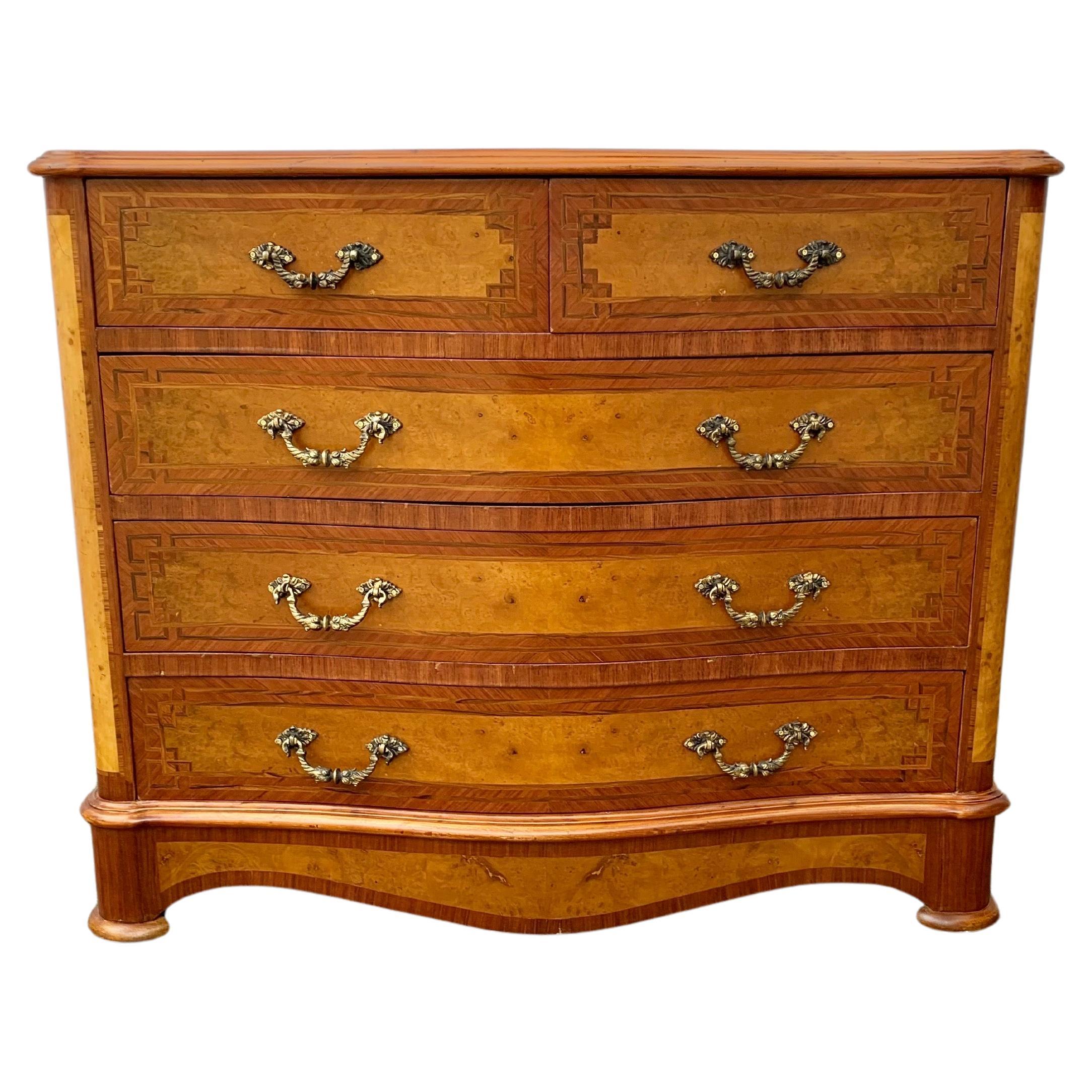 Questa cassettiera o commode del XX secolo è stata realizzata a metà del 1900 con impiallacciatura di radica e noce su una struttura in pino. Il piano è caratterizzato da un piano disegnato in legno che poggia sopra cinque cassetti, due più piccoli