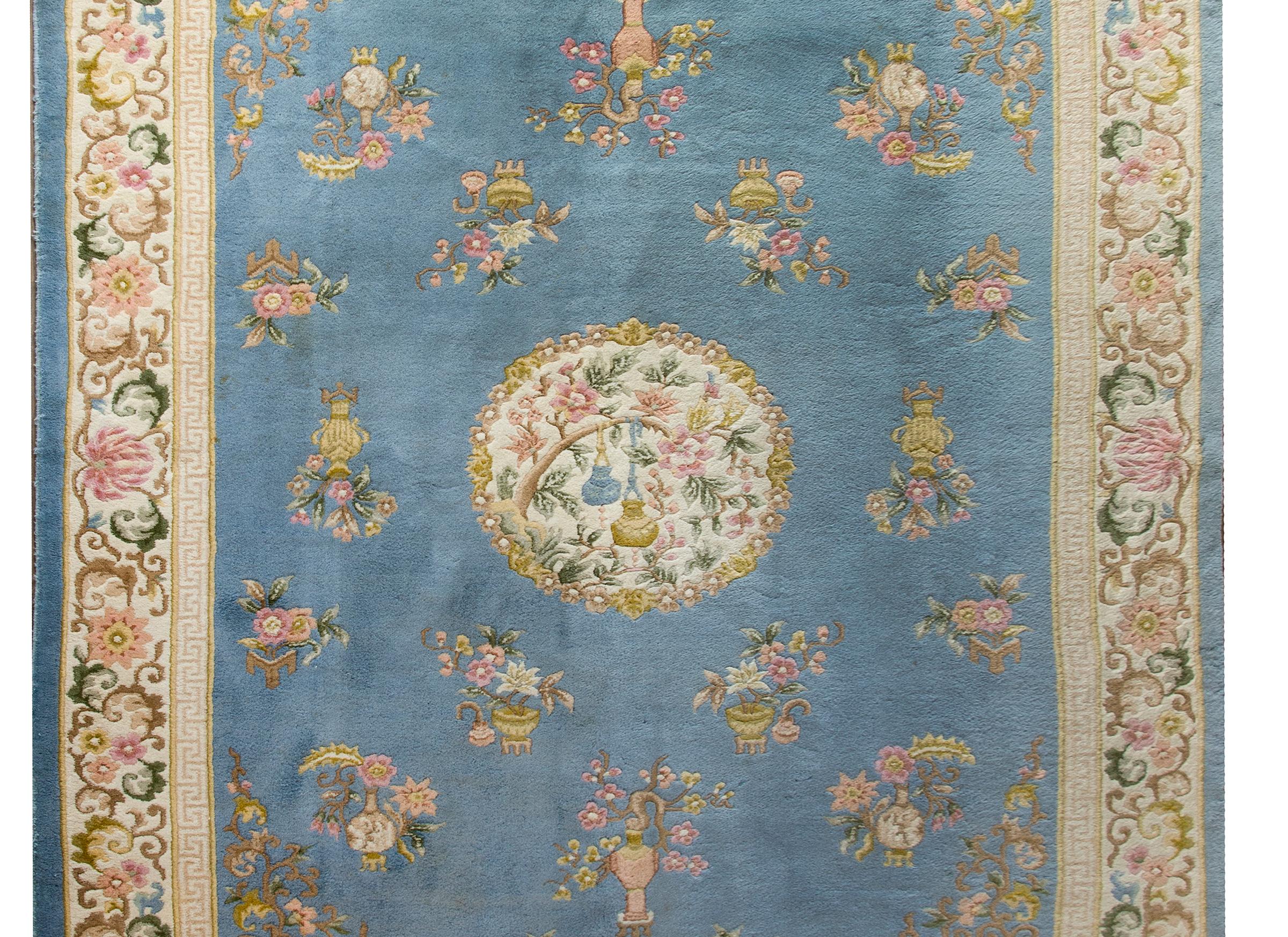 A sweet late 20th century Chinese rug with myriad potted flowers including chrysanthemum, peonies, and prunus blossoms all woven in pale pinks, yellows, greens, and whites, and set against a pale indigo background. The border is wild with