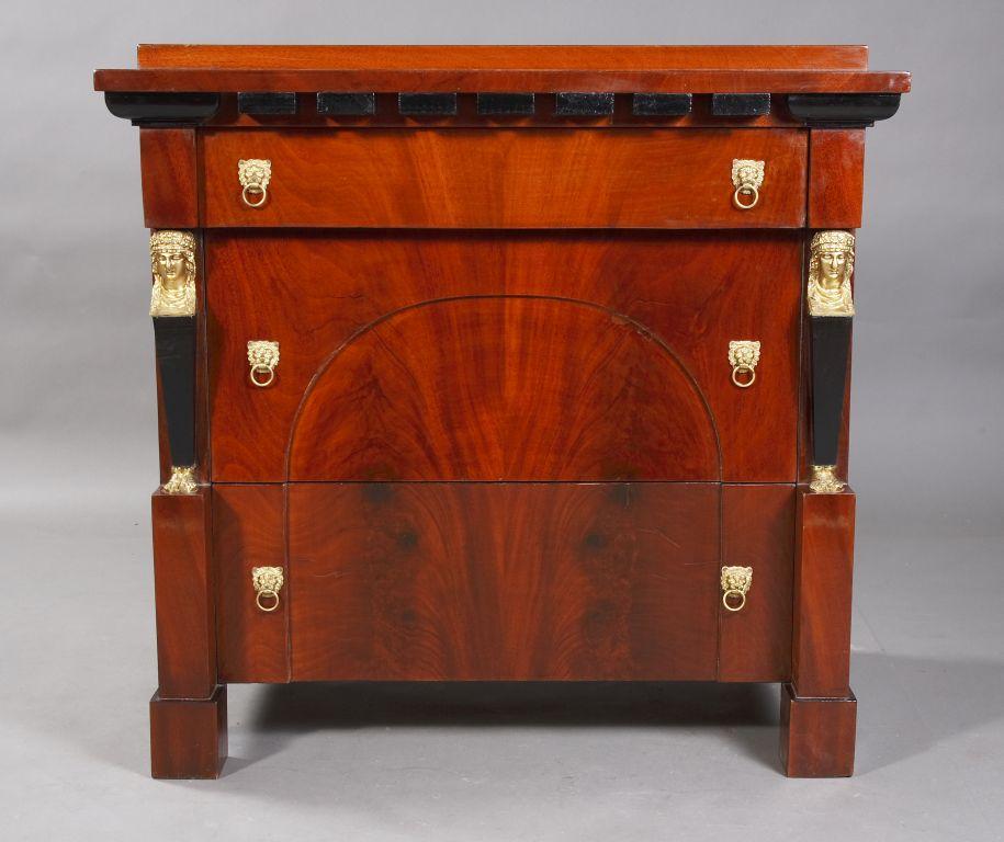 Pyramid mahogany on solid softwood. Straight body on four blocks feet. Architecturally structured front of three drawers of different sizes, below segmental filling, flanked by caryatids. Slightly protruding, profile-framed cover plate. Dental