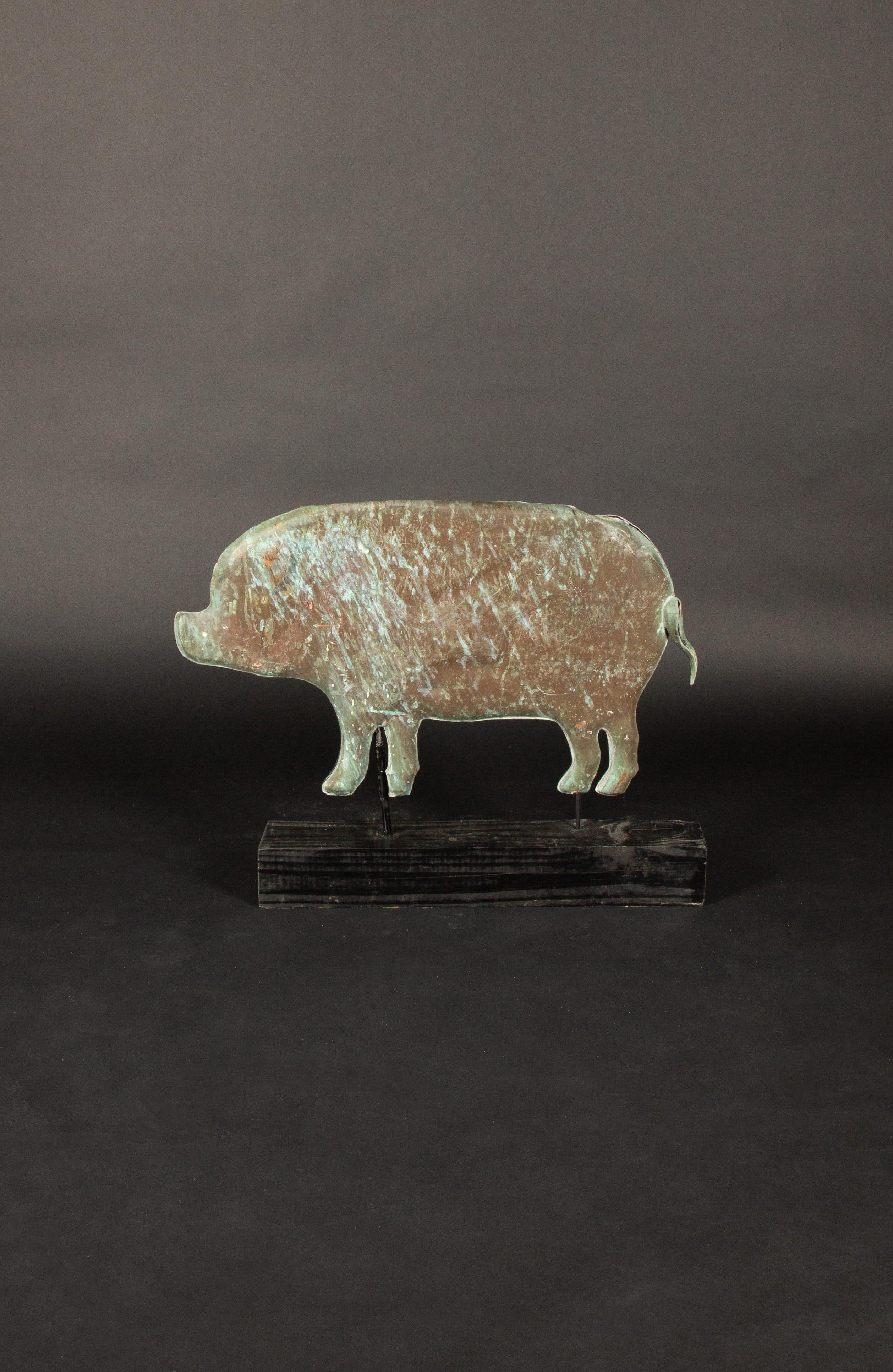 This charming 20th-century copper pig weathervane, now mounted on a sturdy wooden base, showcases a beautiful patina that highlights its aged character. The silhouette of the pig is rendered in a primitive folk-art style, emphasizing its handcrafted