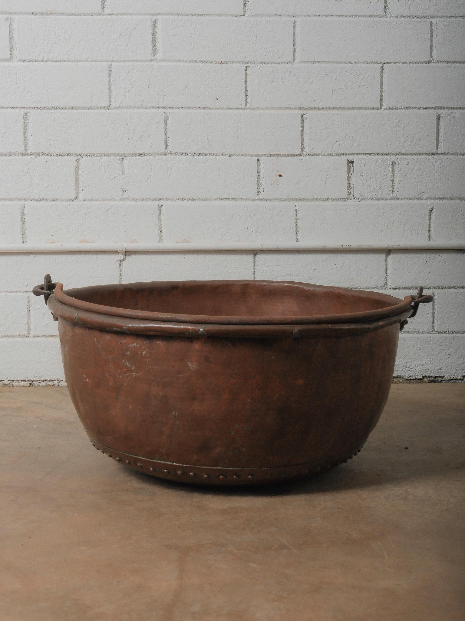 A commanding hand-forged copper vessel defined by its rounded, architectural form and a single iron swinging handle that arcs cleanly across the top. The handle—subtly irregular from its hand-wrought construction—moves fluidly on forged eyelets,