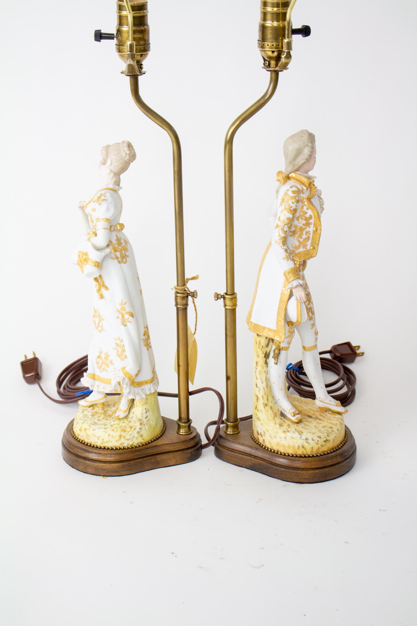 20th Century courting couple bisque figural lamps, a pair. A fancy couple dressed in bright white and gold lean away from each other playfully. She holds a bonnet in her hand, and he holds a cap. Their dress is from the time of Napoleon Bonaparte.