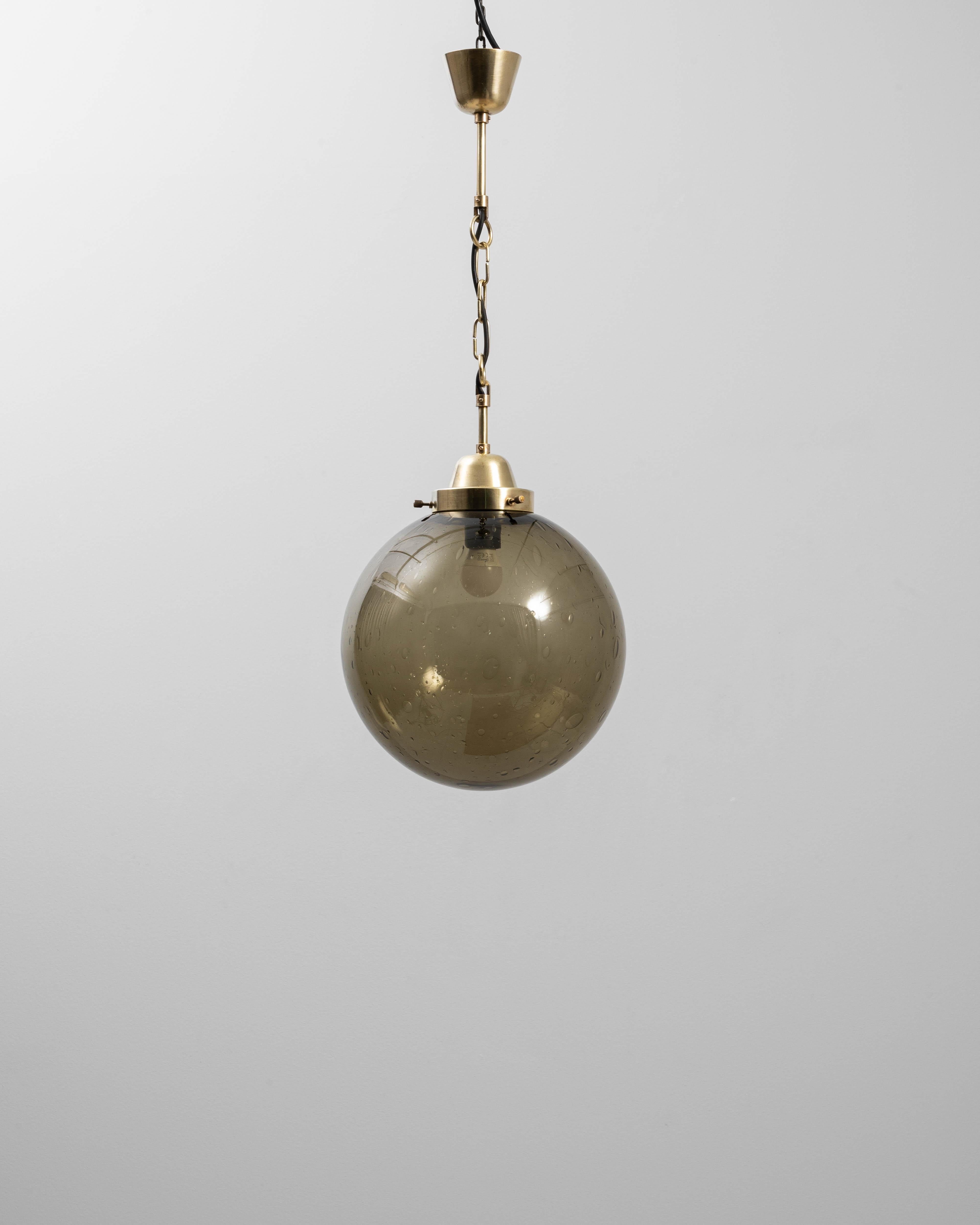 Questa lampada a sospensione ceca del XX secolo presenta una straordinaria combinazione di ottone e vetro fumé, che riflette la raffinata maestria del design europeo di metà secolo. La lampada presenta un paralume sferico in vetro soffiato a mano