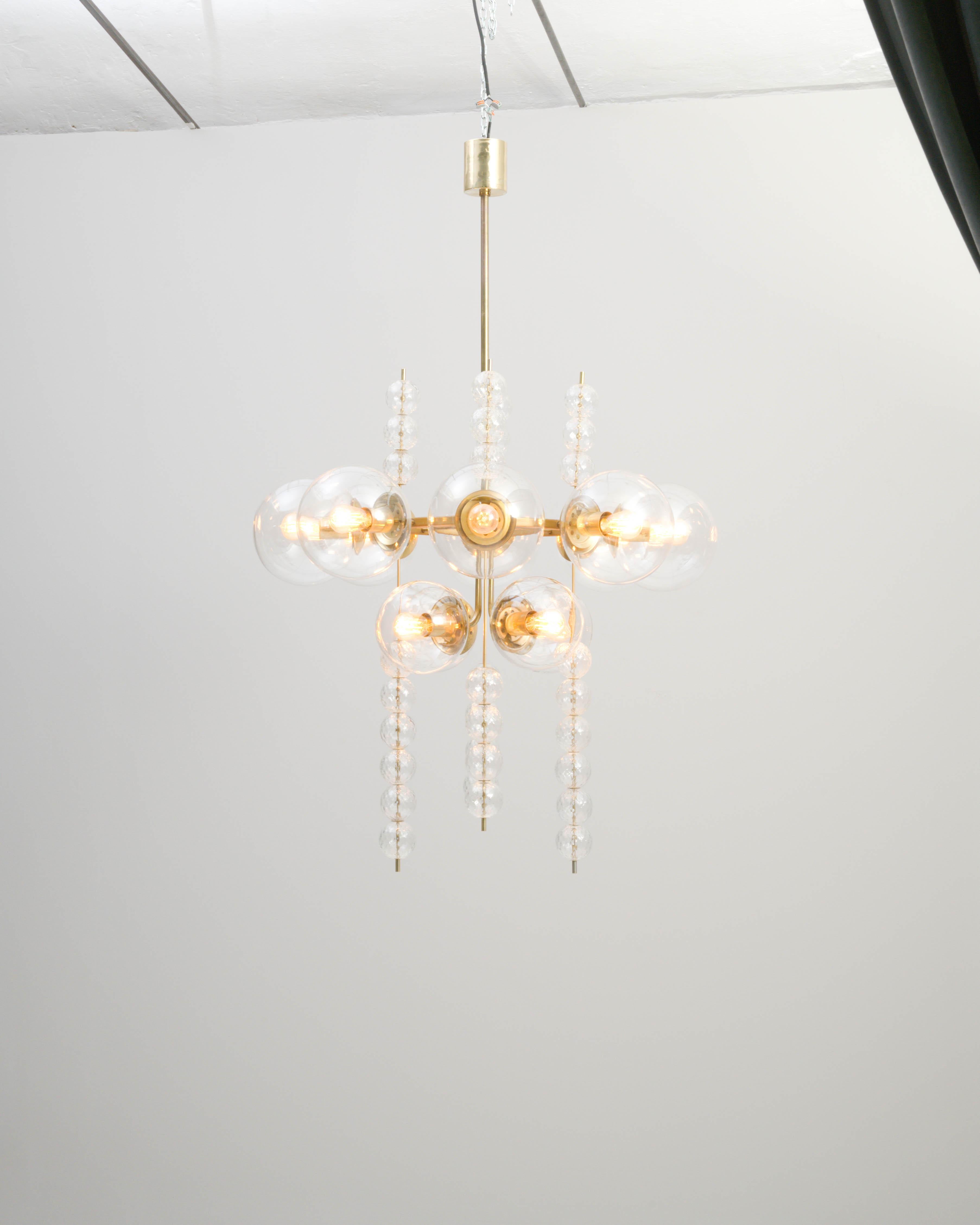 This vintage chandelier conjures an air of timeless glamour. Made in Czechia in the 1970s, glass globes create an impression of ethereal beauty, while the polished brass frame lends a subtle Industrial inflection to the design. Vertical strings of
