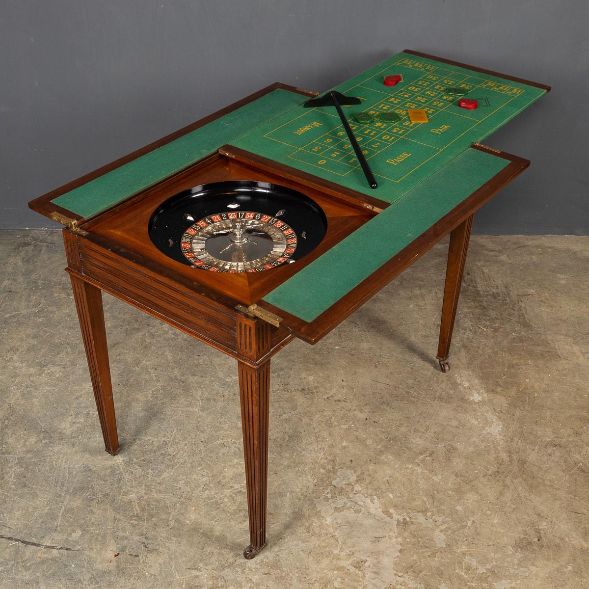 Antique early 20th century Edwardian gaming table in rich mahogany, the top of the table opens to reveal upholstered fine green felt fabric surface, set with a roulette wheel, dice and two shakers, gambling chips, cribbage board and playing cards.