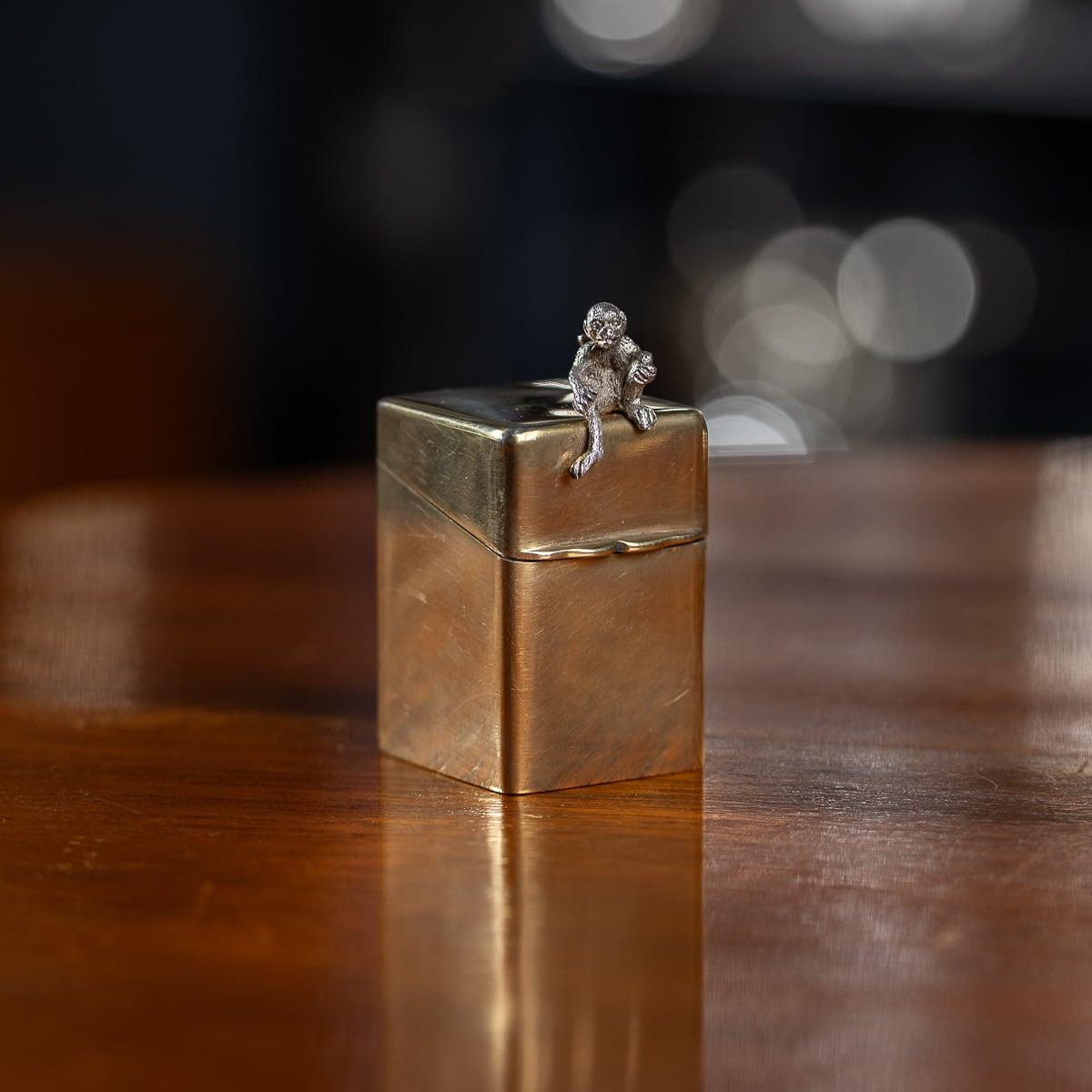 Antigua caja inglesa del siglo XX para frascos de perfume de plata maciza chapada en oro de 22 quilates, diseñada con forma cuadrada y tapa de apertura en ángulo. Un mono plateado se posa sobre la tapa. En el interior, la caja contiene dos pequeños