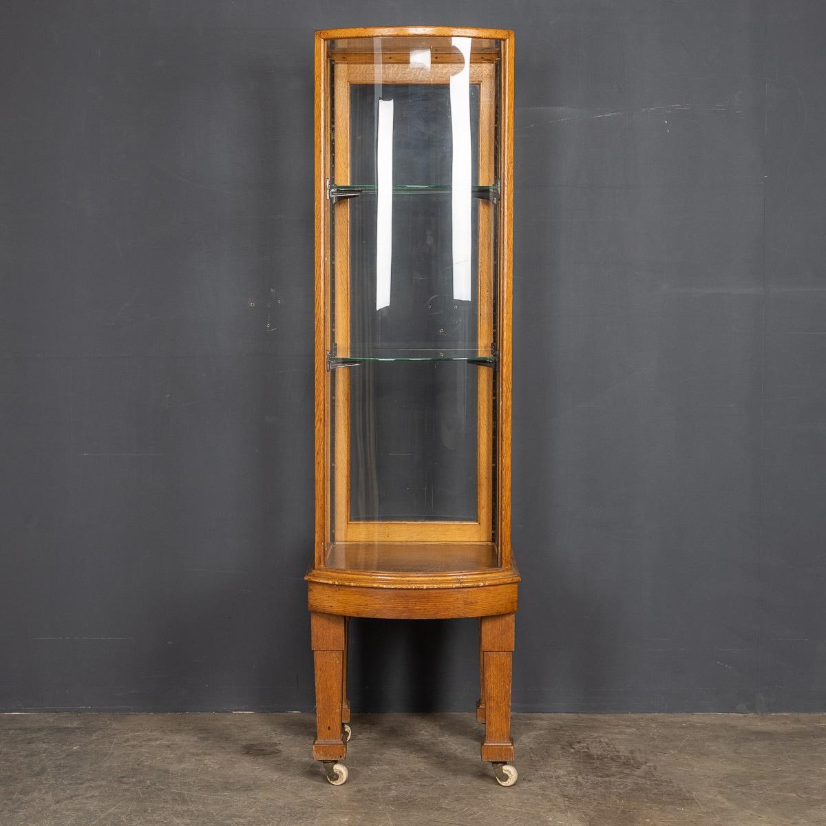 Antique 20th century English Art Deco slender oak and glass display cabinet with two adjustable glass shelves. With a glass bow front and a rear door with original latch, standing on solid square legs ending in original castors, a very practical and