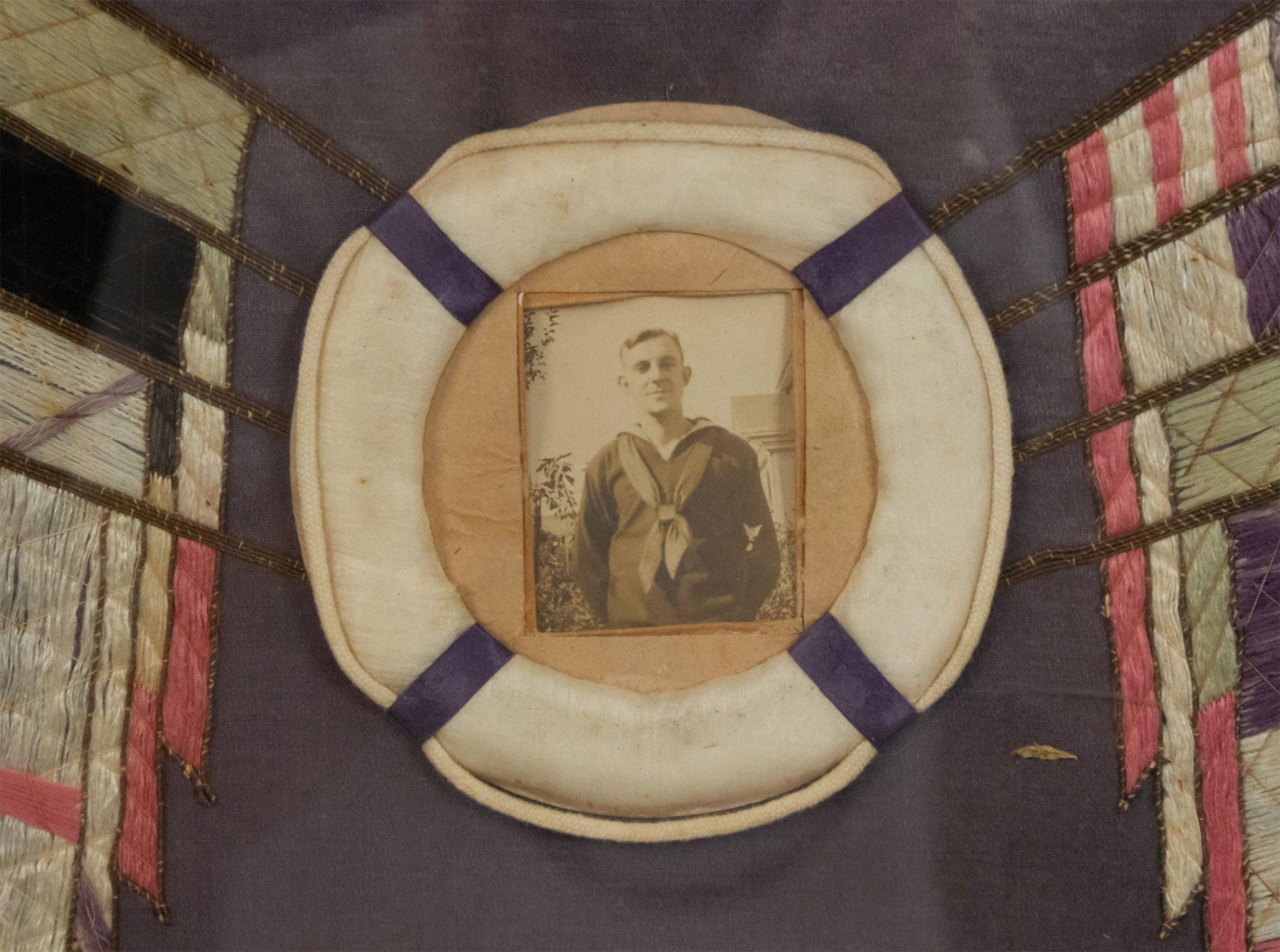 English (20th century) ebonized framed memorial embroidery of flags and ships with photograph of a sailor in a life raft. Text embroidered at top, 