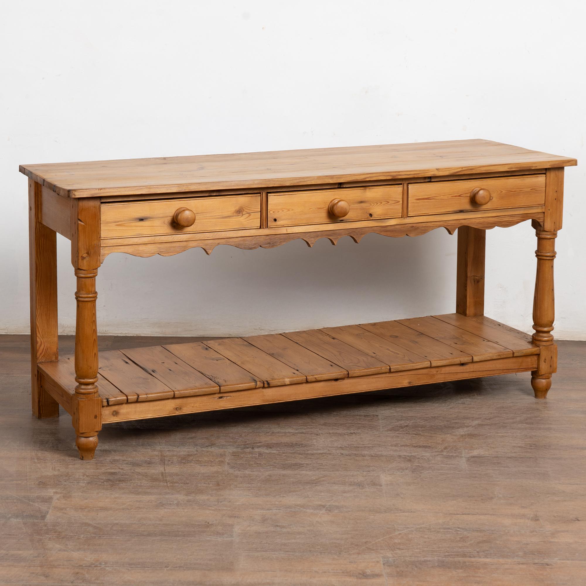Console en pin anglais du 20e siècle comprenant trois tiroirs avec de grandes poignées en bois, une jupe festonnée, des pieds tournés et une étagère inférieure.
Restaurée, solide et stable, elle est idéale comme console, table de canapé ou îlot de
