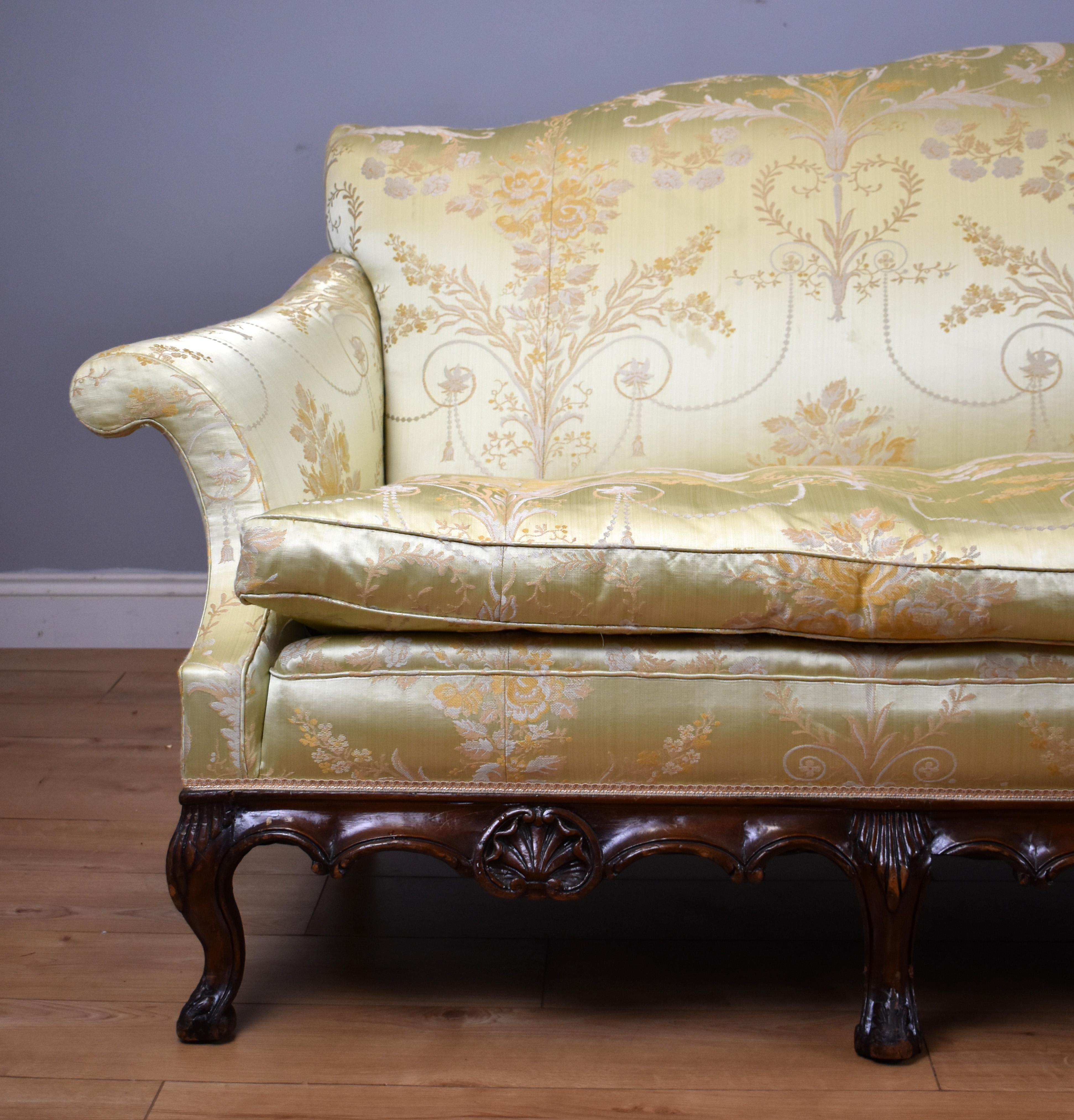A Queen Anne style sofa, upholstered in pale yellow foliate patterned silk type material, with exposed shell and scroll carved frame raised on four front cabriole legs. The sofa is in good useable, showing minor wear commensurate with age and use.
