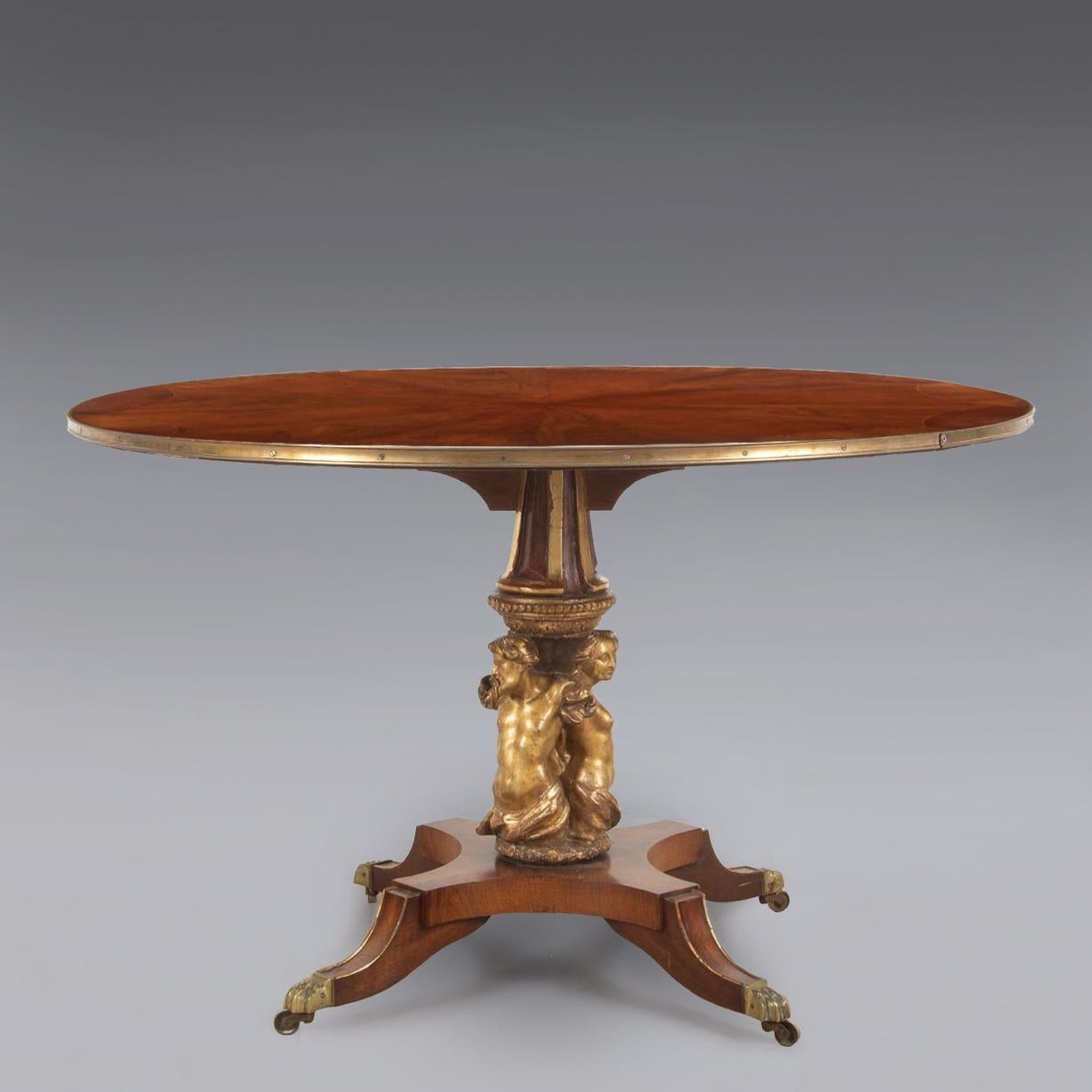 A highly unusual English breakfast or tilt-top table with a radially veneered circular top of exotic timbers bound in brass. 

The giltwood column with beautifully carved classical male and female busts sitting on a quatrefoil base raised on sabre