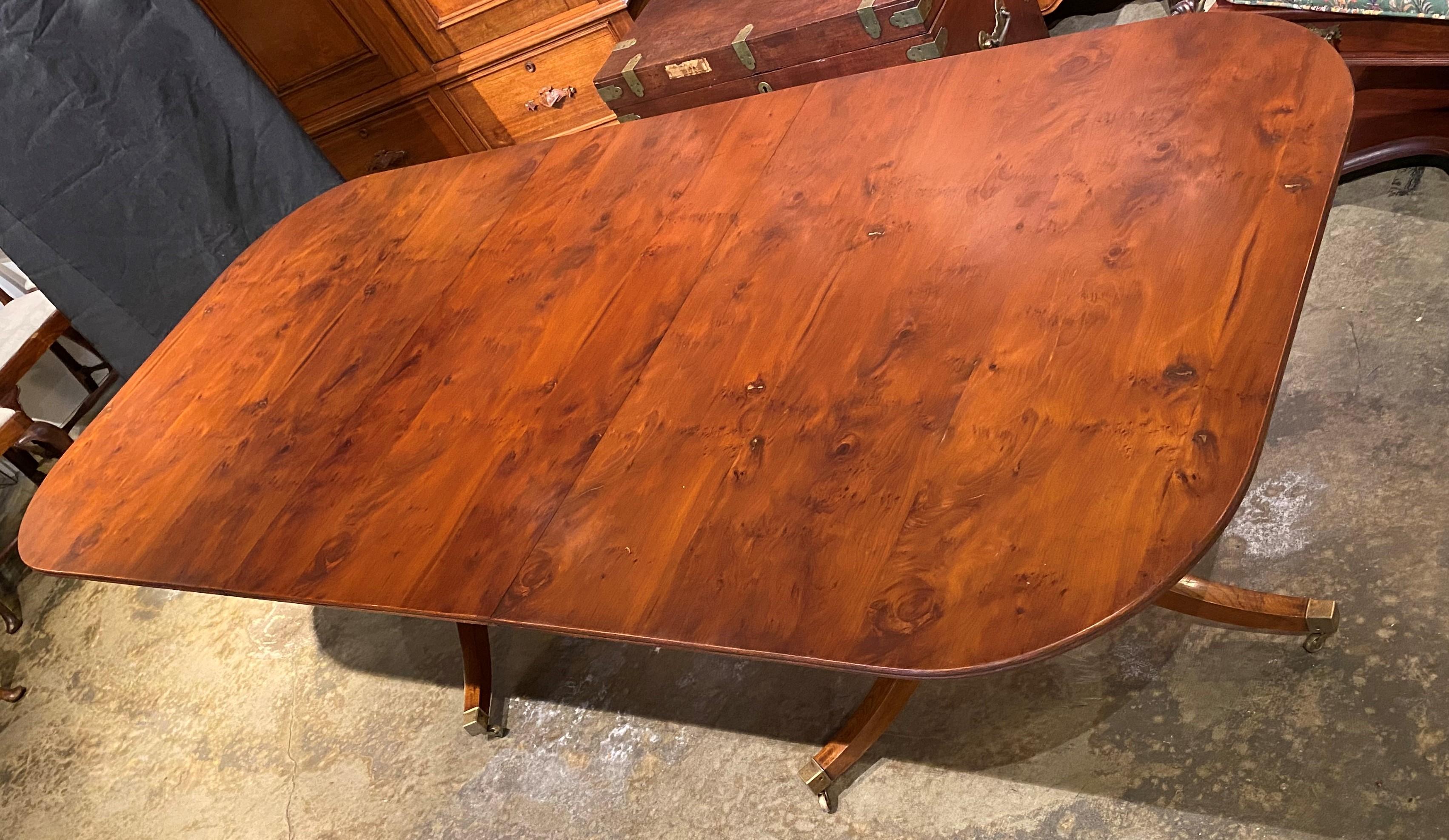 A fine example of a double pedestal dining table with yew wood top with tilt top sections with out swept tripod bases terminating with brass caps and casters, expandable with one large 27.75 inch leaf. English in origin, dating to the 20th century