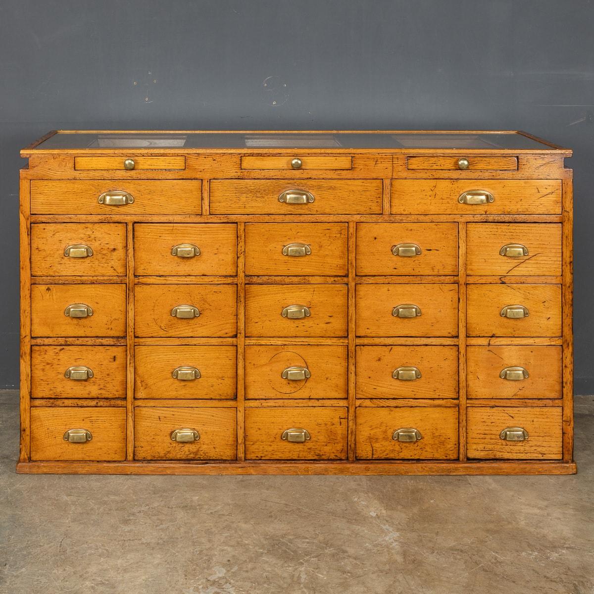 Antique 20th Century English oak haberdashery counter has a glass top showing three display drawers, under these are three wide slim drawers, beneath these are twenty drawers all have their original brass handles and knobs. In the front of the