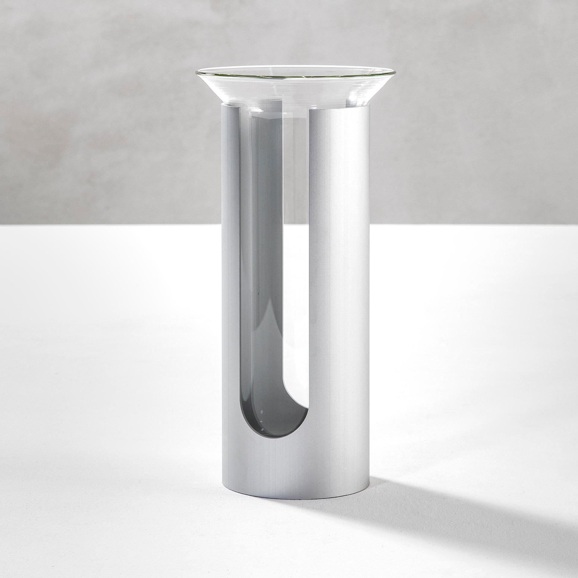 Camicia was designed by Enzo Mari in 1961. Camicia is a vase that consists of a double element: a cylinder of opaque anodized aluminum, bottomless, which supports and envelops a transparent glass container. The hole and the full-height vertical cut