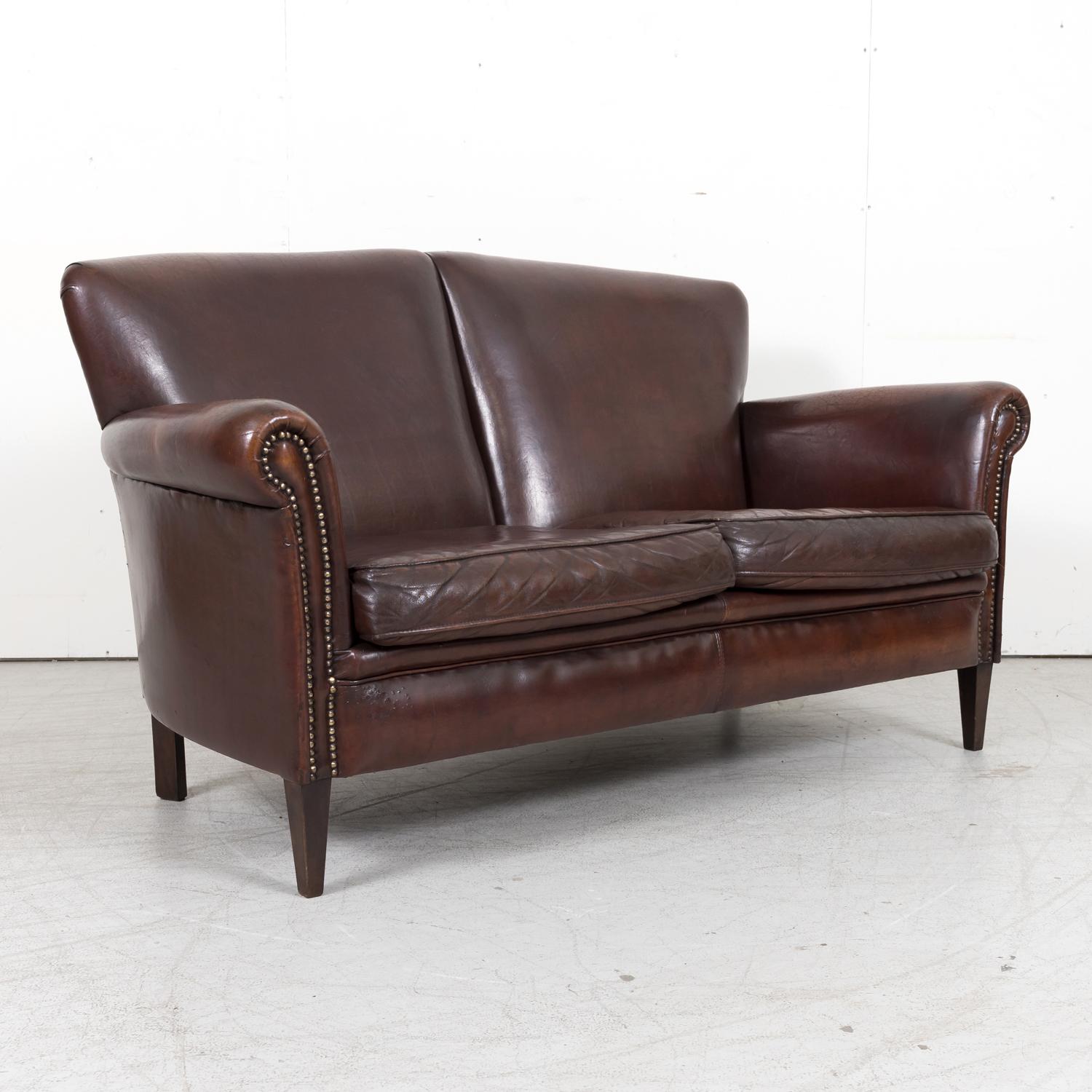A wonderful 20th century French Art Deco two seater settee or sofa in original dark brown leather, circa 1930s, having a tight back, scrolled arms with nailhead trim, and two loose seat cushions. Raised on short tapered walnut legs. All original.