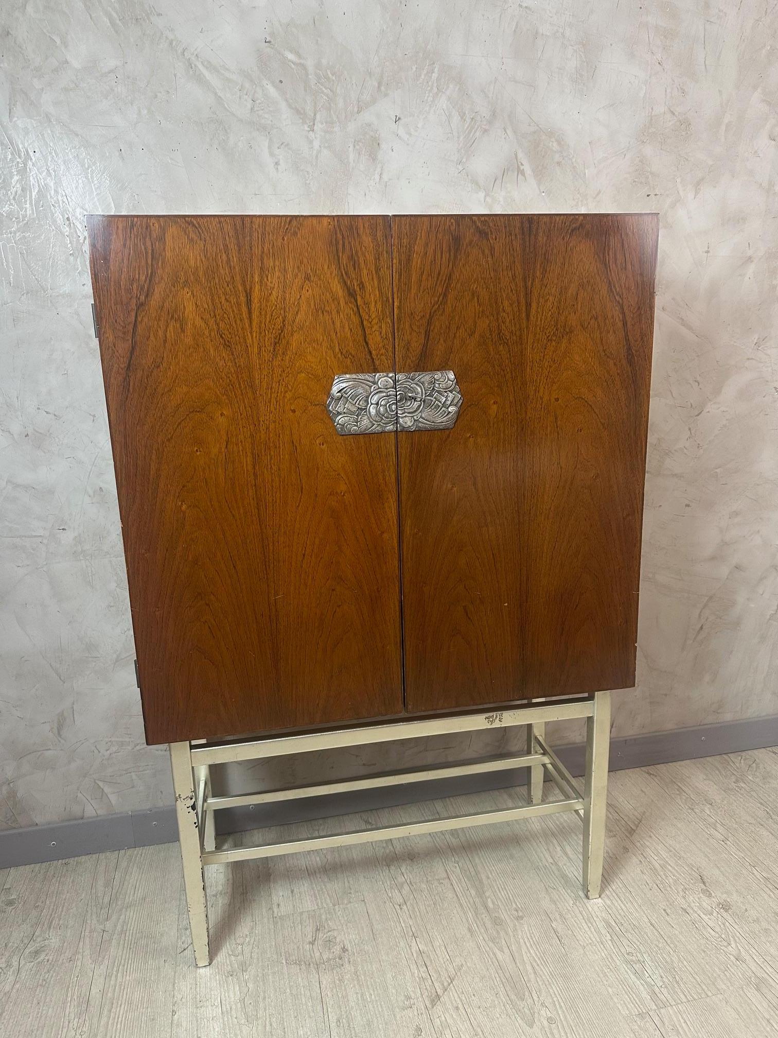 Elegante mueble bar Art Decó que combina la calidez de la chapa de nogal con las elegantes líneas de una base de metal plateado.
Las puertas dobles se abren para revelar un refinado interior equipado con estantes de cristal, parte trasera de espejo