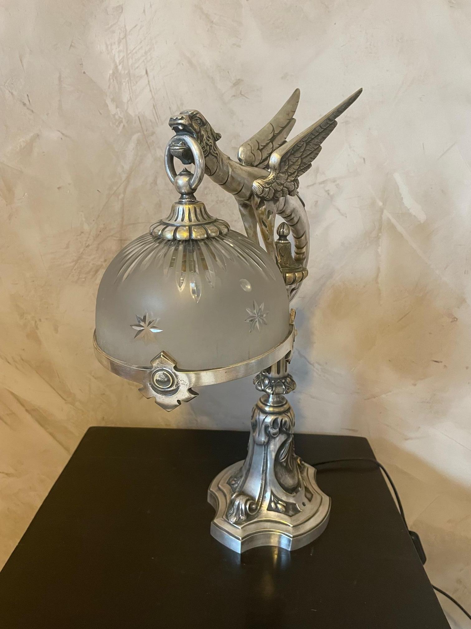 Exceptionnelle lampe de table d'époque art nouveau en bronze argenté, représentant une chimère tenant dans sa bouche le verre ciselé de l'abat-jour. 
L'abat-jour est entouré d'une couronne de bronze. 
Nous avons électrifié la lampe. 
Belle