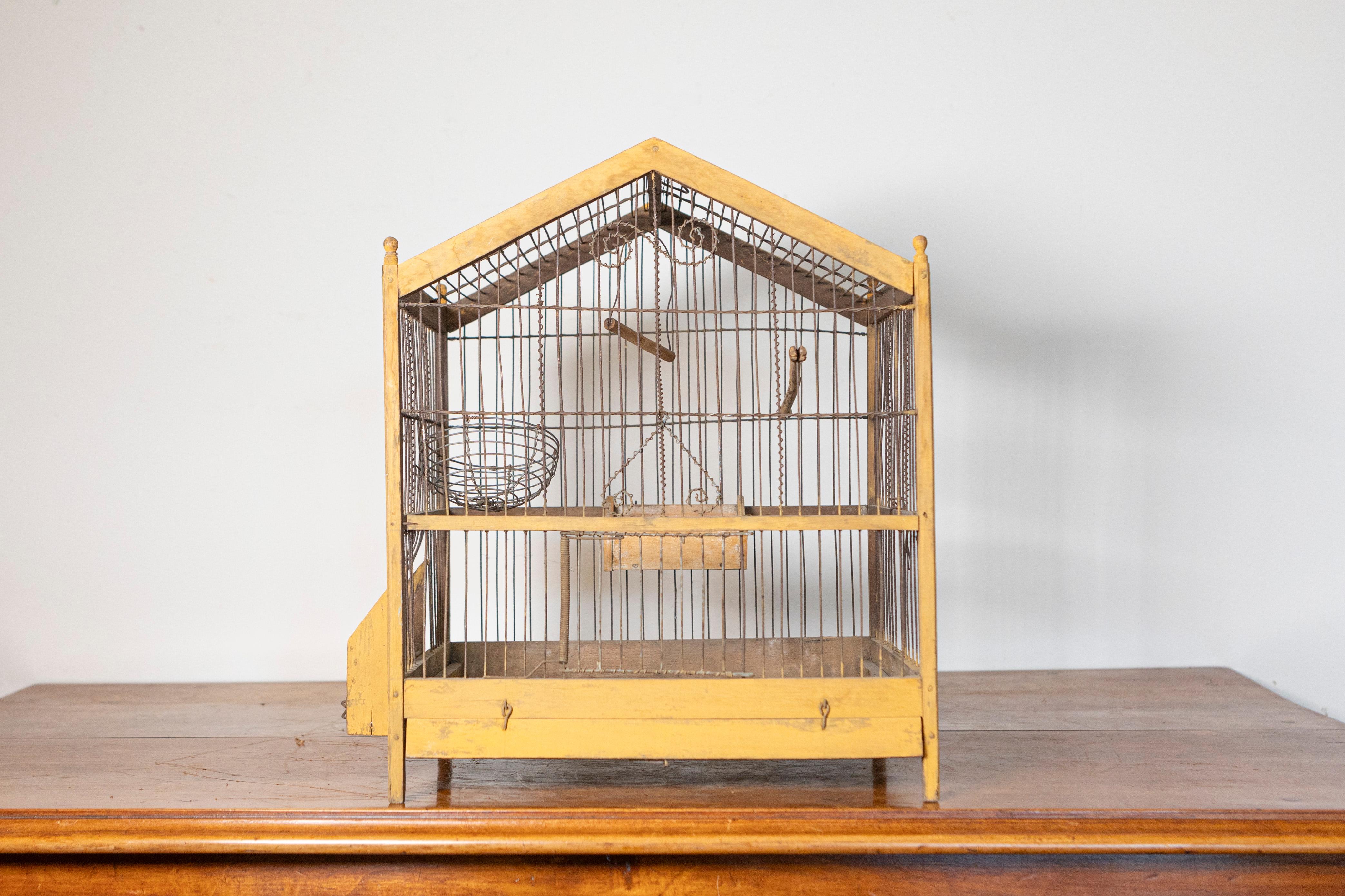 This charming early 20th-century French birdcage is crafted from painted wood and iron, featuring a house-shaped structure with a pitched roof. The soft yellow-painted wooden frame adds warmth and character, complementing the delicately spaced iron