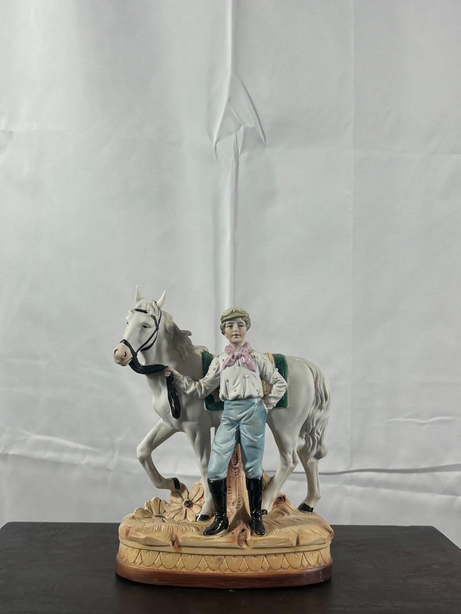Jolie figurine en biscuit (mélange de porcelaine et kaolin) datant des années 20 représentant un cheval et son cavalier habillé d'une chemise blanche avec des perles dorées, un jean, un foulard rose, une casquette et des bottes noires vernissées.
