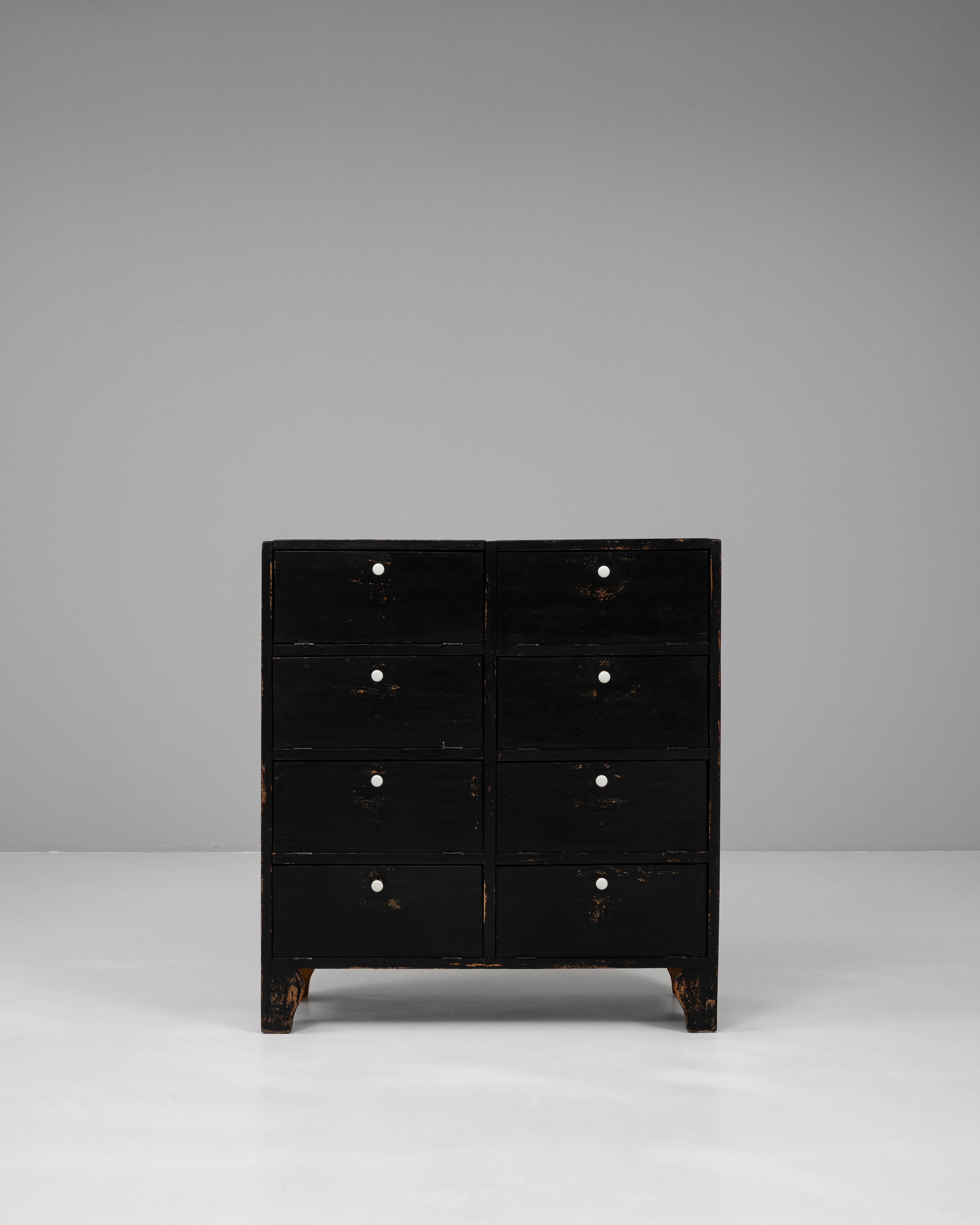 Questa cassettiera in legno patinato nero francese del XX secolo unisce il fascino dell'utilità a un'estetica sorprendente e fuori dal tempo. La sua forma compatta ospita otto cassetti frontali, ognuno dei quali è rifinito con una superficie nera e