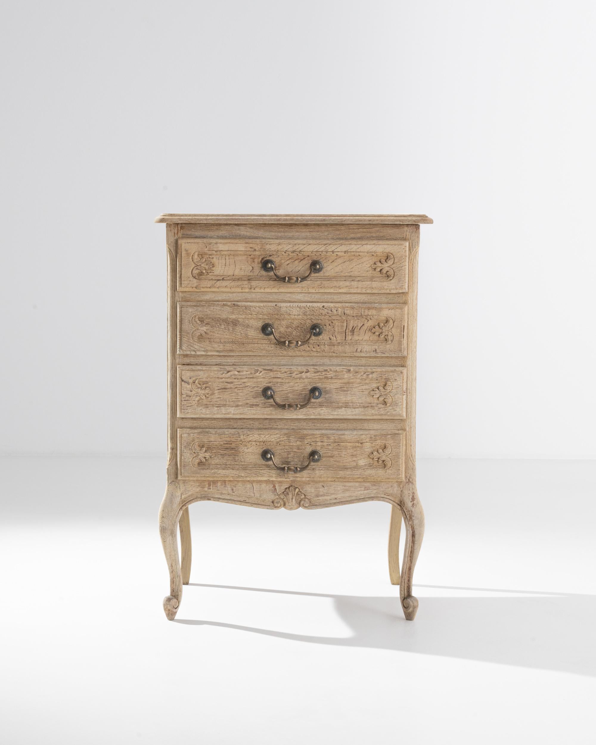A 20th century bleached oak chest of drawers made in France. Four top-down drawers flanked by animatedly curved legs shape this handsome set of drawers. Scroll and leaf-patterned enlays, combined with sharp metal drawer handles, craft a dutiful