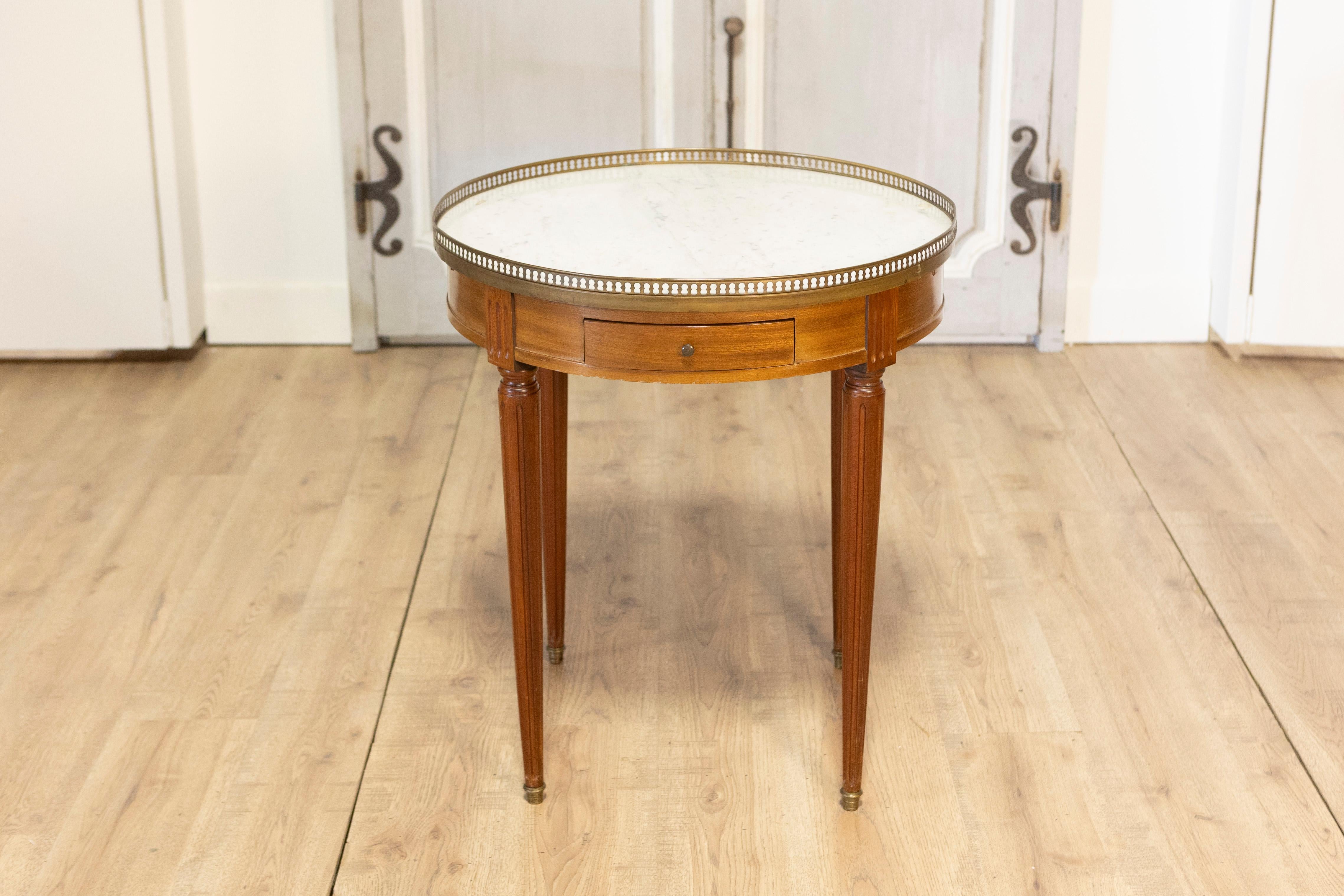 This 20th-century French Bouillotte table exemplifies the timeless elegance of neoclassical design. Crafted from mahogany, the table features a circular marble top surrounded by a pierced brass gallery, combining functionality with refined