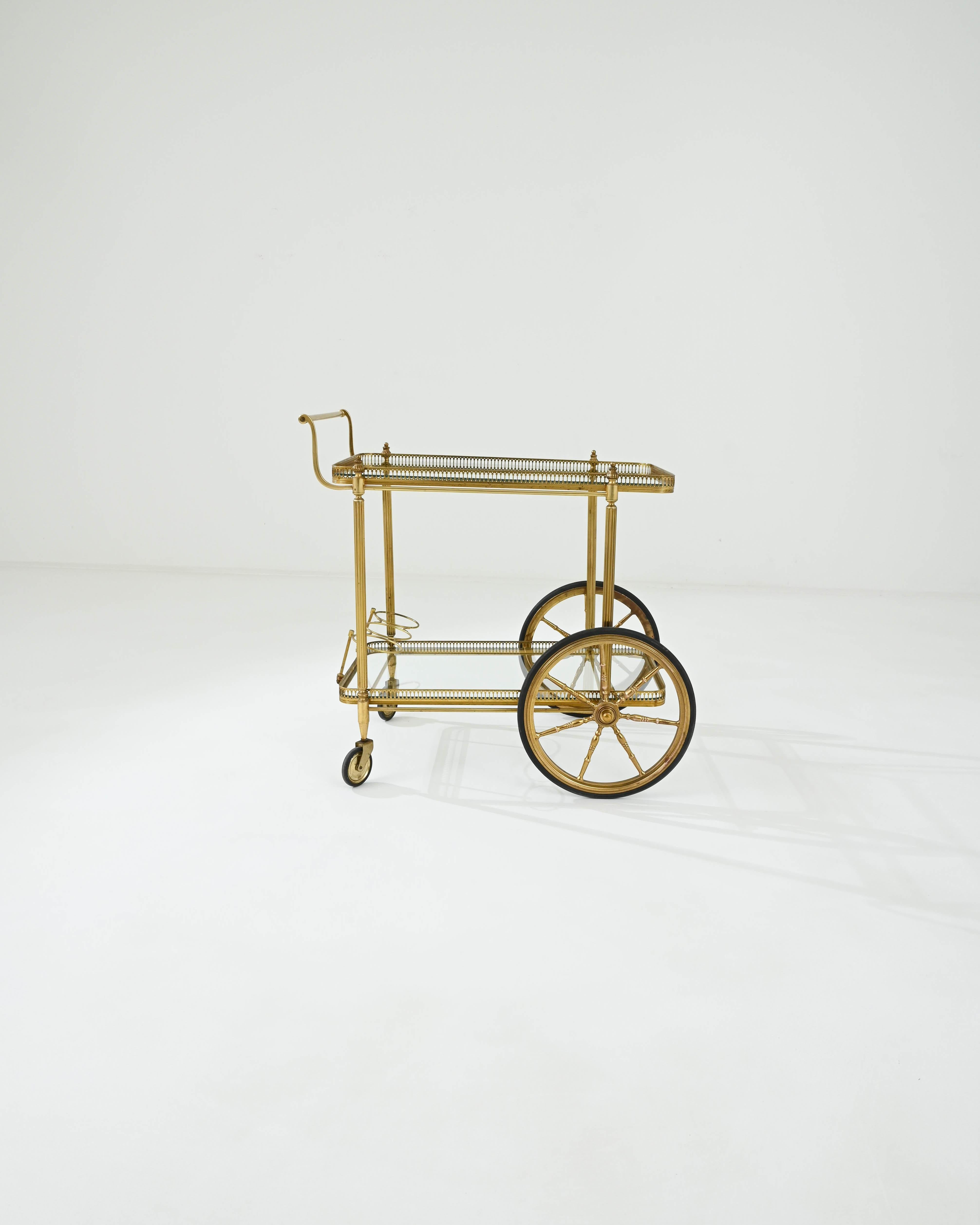 A brass bar cart created in 20th century France. Created in art deco fashion, this gleaming bar cart shines with a brilliant sense of luxury and opulence. An elegant composition is created by the detailed legs, the intricate small fence that