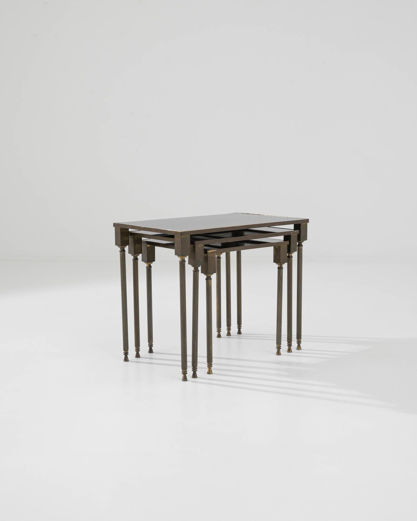 This trio of nesting tables provides a sophisticated accent. Made in France in the 1960s, a gilded brass base, gold-colored legs, and darkly tinted glass form a pleasing color palette. The geometric legs give a modern inflection to the Neo-classical