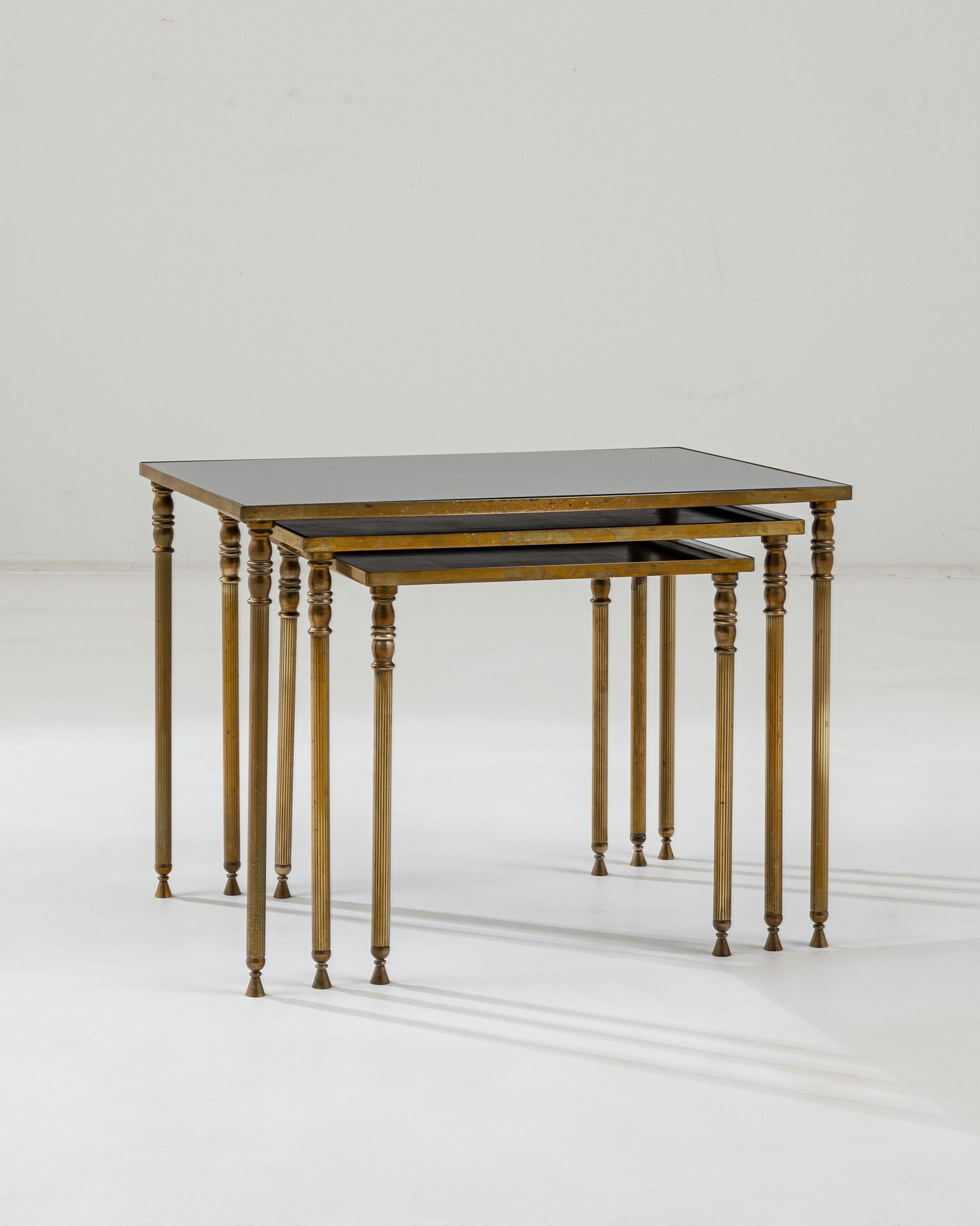 This trio of nesting tables provides a sophisticated accent. Made in France in the 1960s, a gilded brass base, gold-colored legs, and darkly tinted glass form a pleasing color palette. Fluted legs, crowned with golden urns, create a Neo-classical