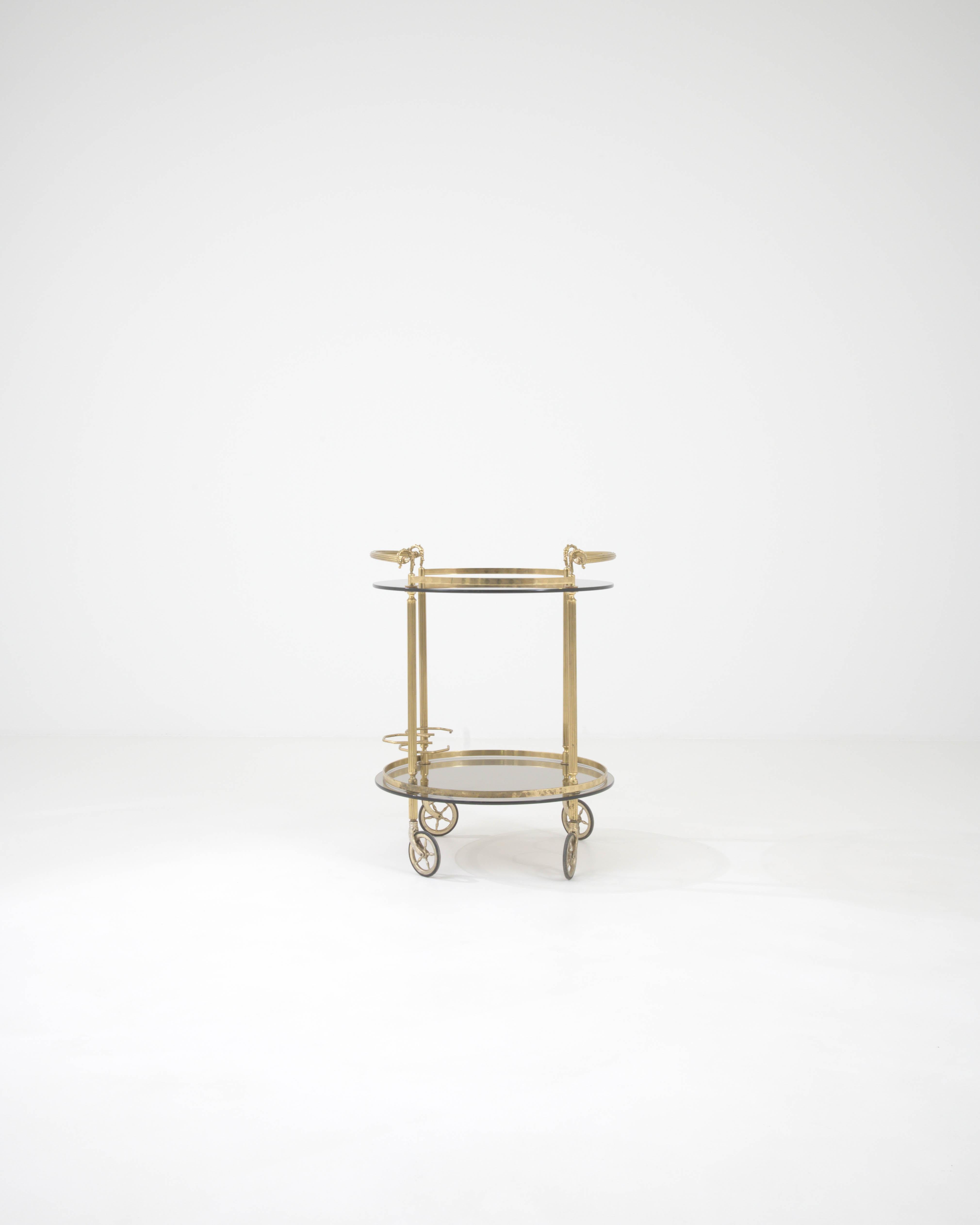 Indulge in the lavish charm of this 20th Century French Brass & Glass Bar Cart, a chic and versatile addition to any modern home. The polished brass structure shines with a timeless glow, accentuated by ornate details such as the elegant finial