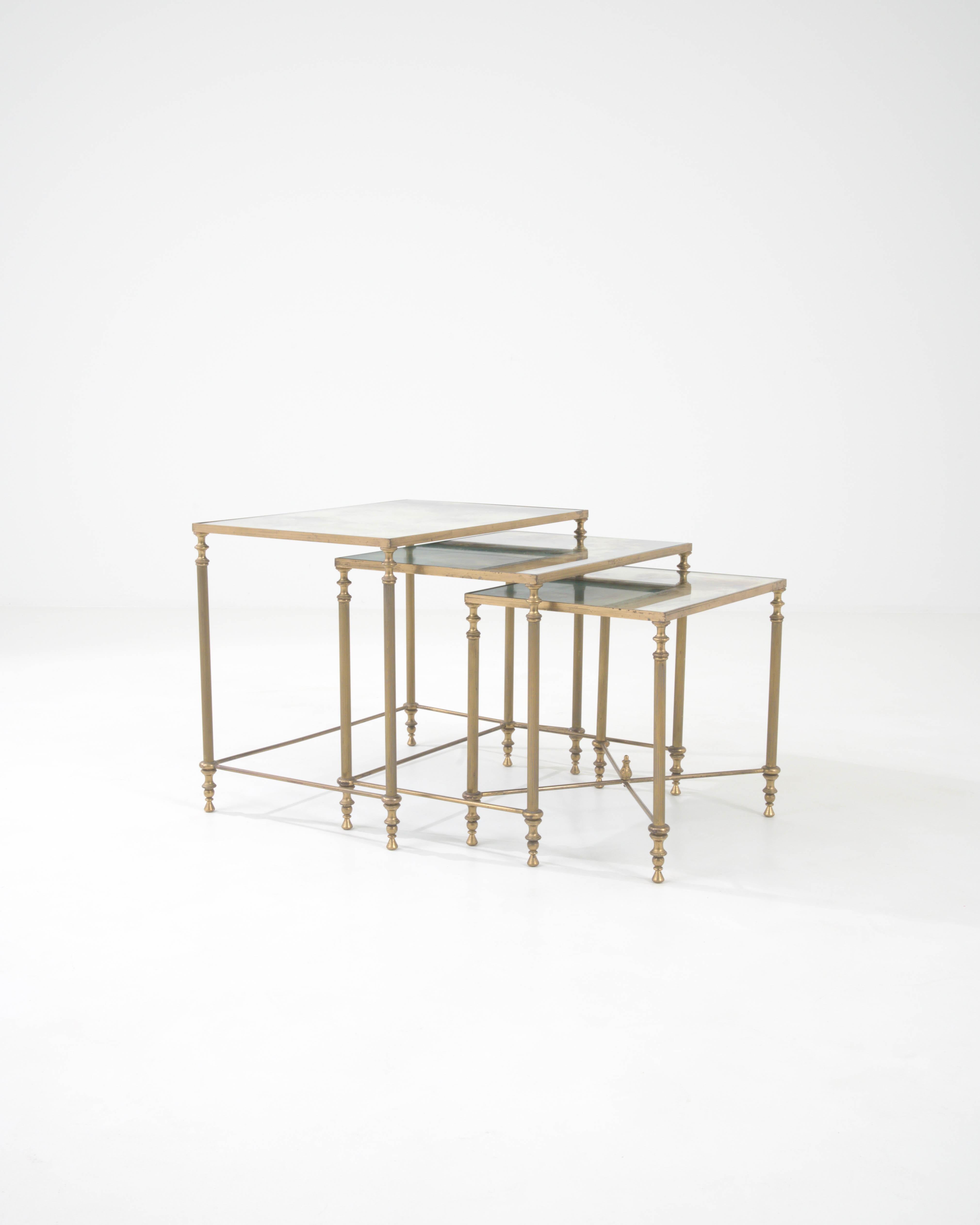 The graceful silhouette of this trio of vintage brass nesting tables makes an elegant impression. Built in France in the 20th century, a rectangular tabletop of tinted glass is clasped within a brass frame. Geometric facets decorate the columnar
