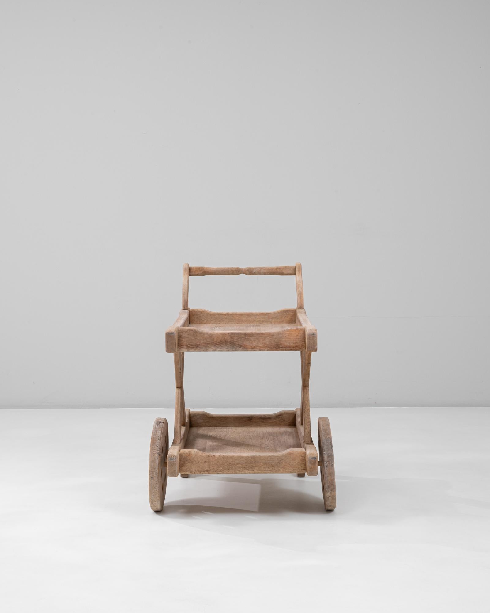 This 20th Century French carved oak bar cart showcases remarkable craftsmanship with its sculpted lines and hand-carved detailing. The piece features two tiers with raised edges and an elegant handle design, supported by an X-shaped frame and large