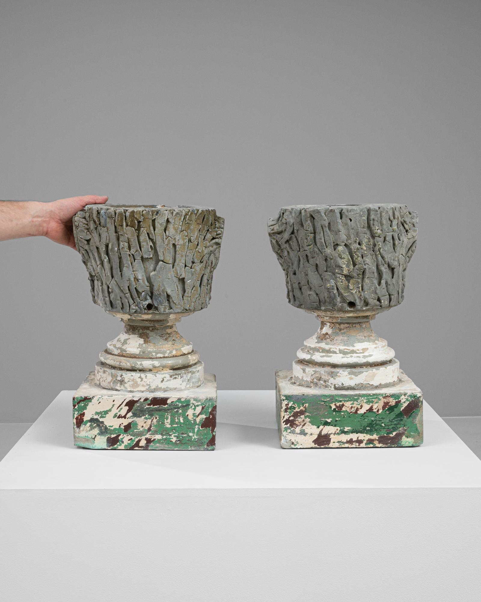 This captivating pair of 20th-century French concrete planters offers a unique aesthetic charm with their heavily textured surfaces and distressed, peeling paint. Each planter stands on a distinctively ornate pedestal base, which contrasts